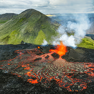 A mountainous landscape is divided in two, with the area closer to the viewer covered in black rock and glowing flows of lava and the area farther from the viewer looking verdant and unaffected by the ongoing eruption.