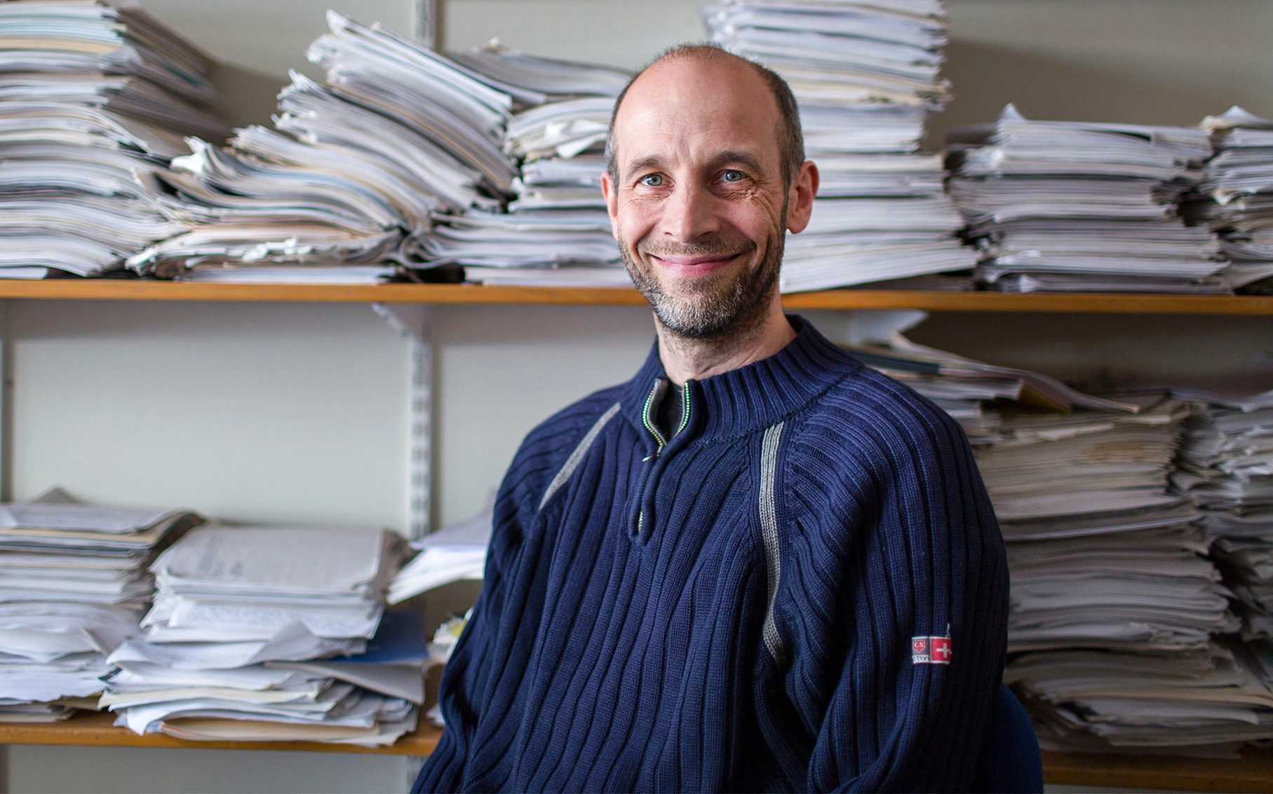Man in a blue sweater smiling in front of piles of papers.