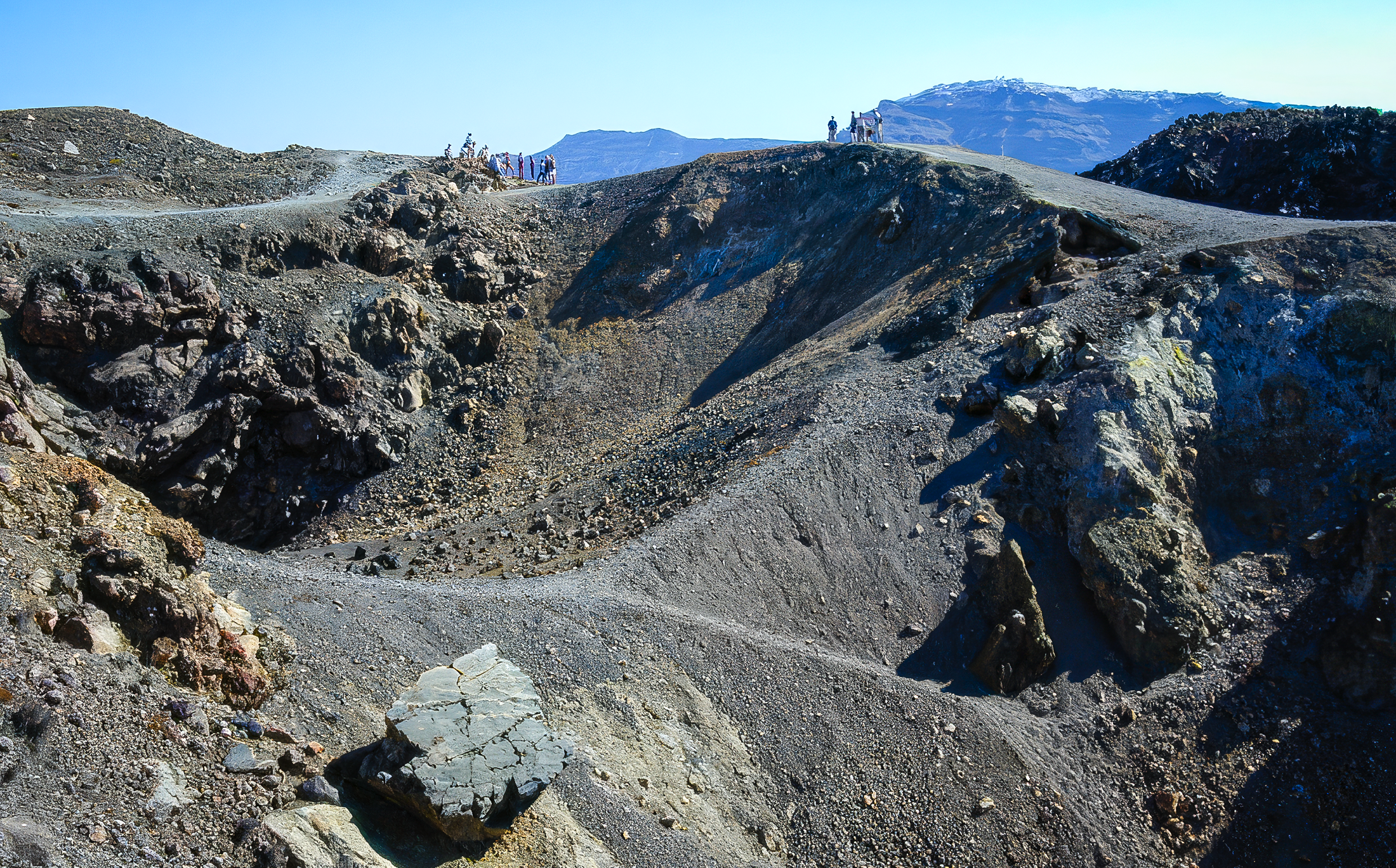 About 20 people, divided into two groups, are visible in the distance, hiking across the top of an ashy volcanic crater.