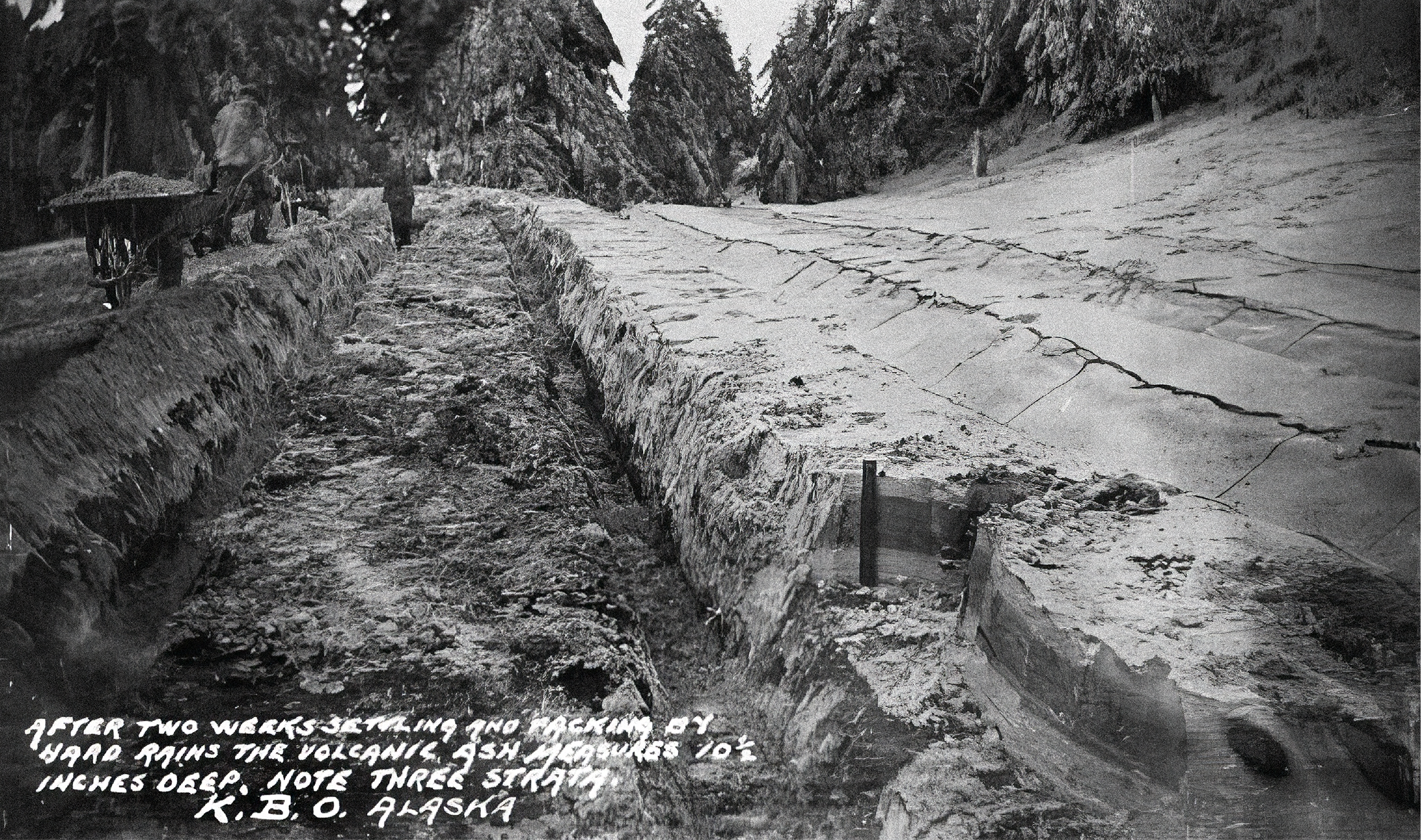 A black and white historical photograph shows people walking along a road covered in deep piles of ash.