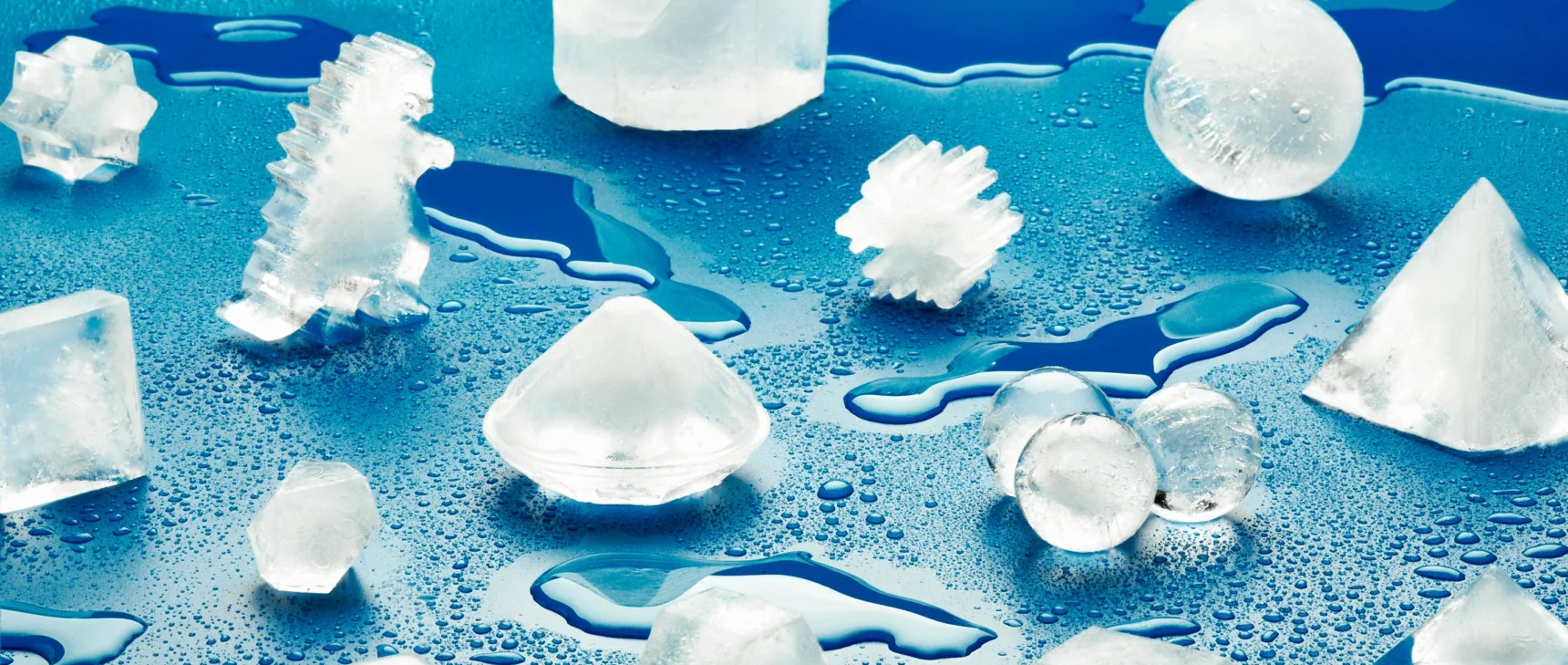 A photograph of ice cubes molded into different shapes—including a sphere, a snowflake, a diamond, and a dinosaur—scattered across a blue surface.
