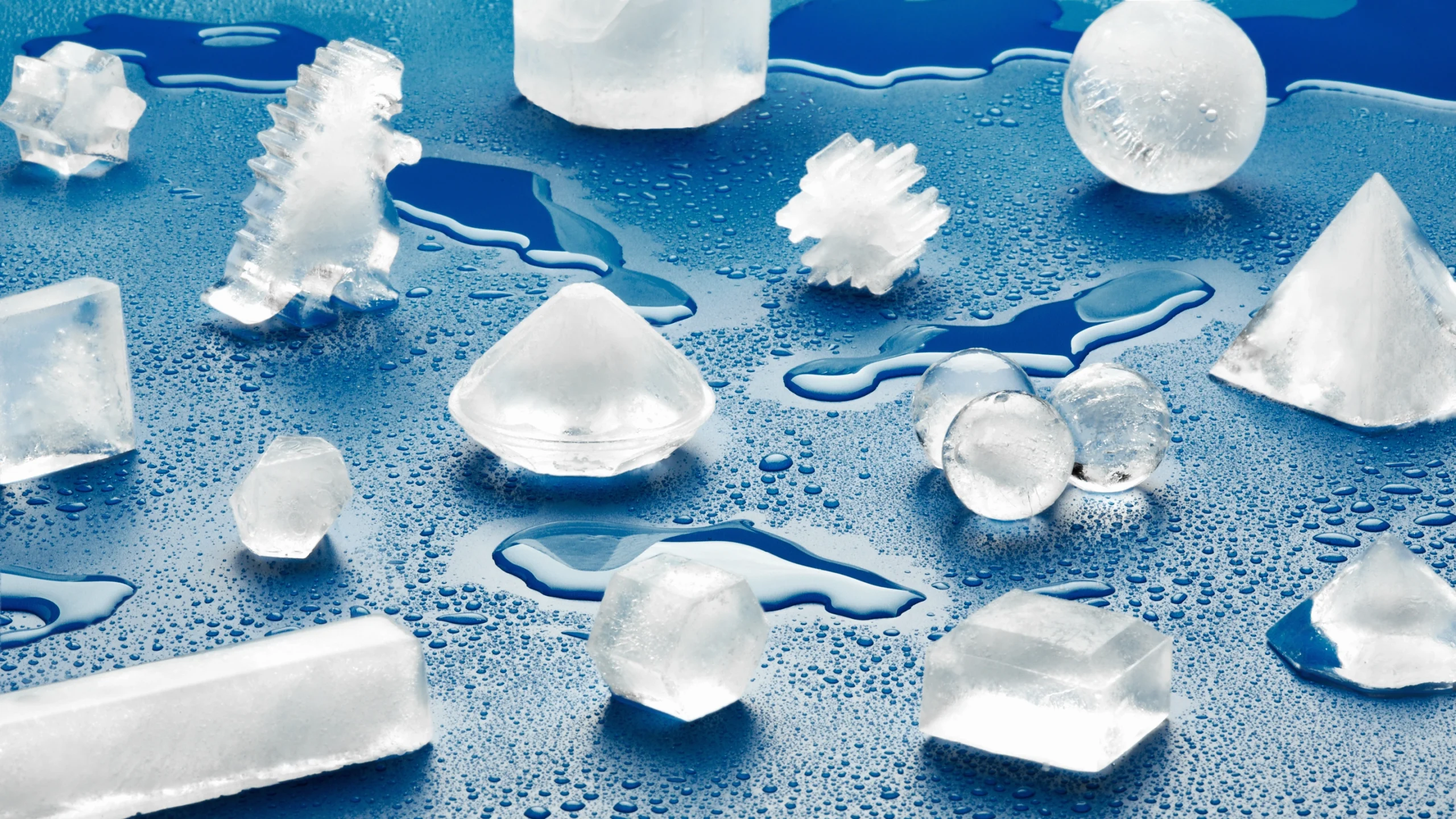 A photograph of ice cubes molded into different shapes—including a sphere, a snowflake, a diamond, and a dinosaur—scattered across a blue surface.