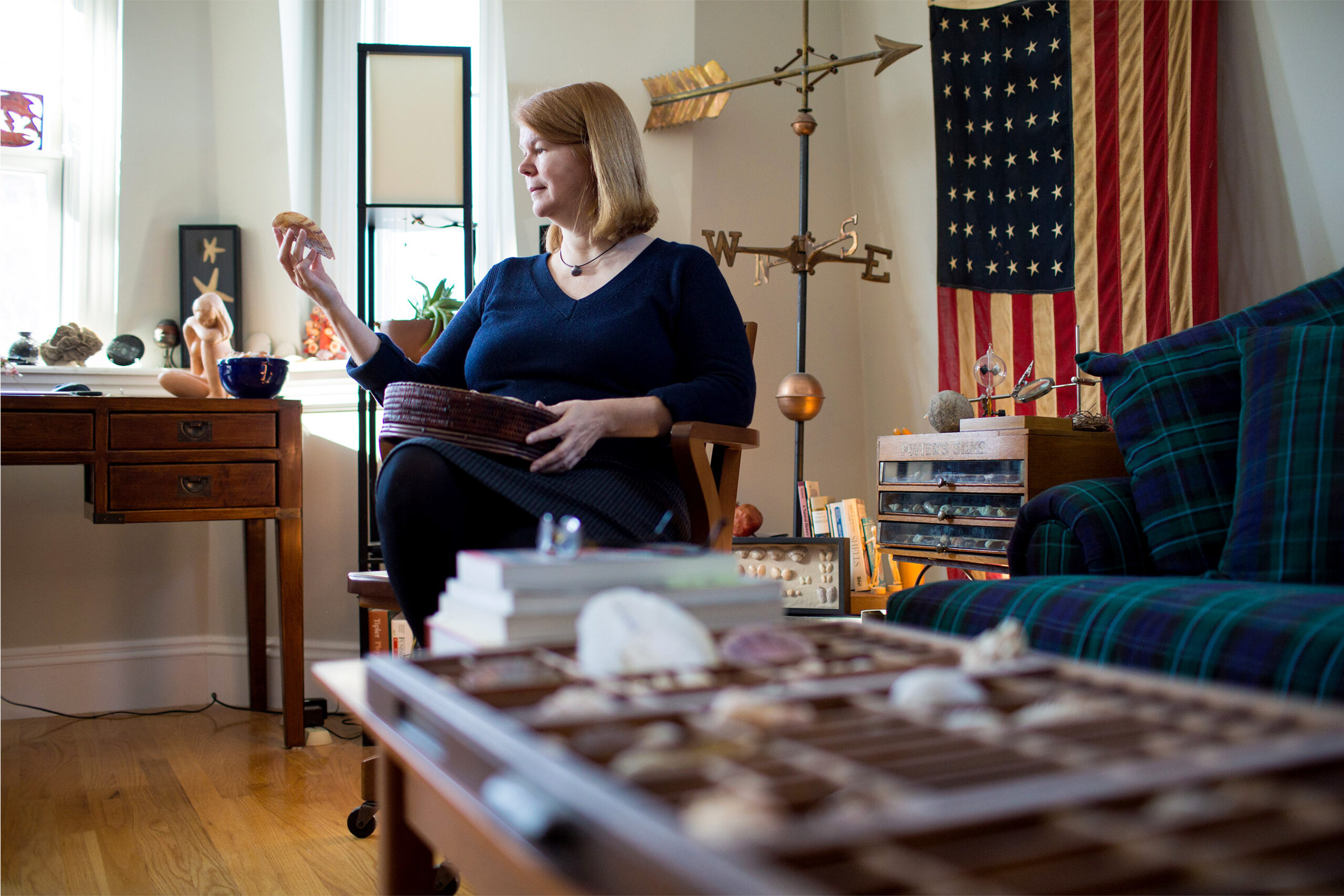 A woman wearing a blue sweater sits in a chair surrounded by a weathervane and other curios.