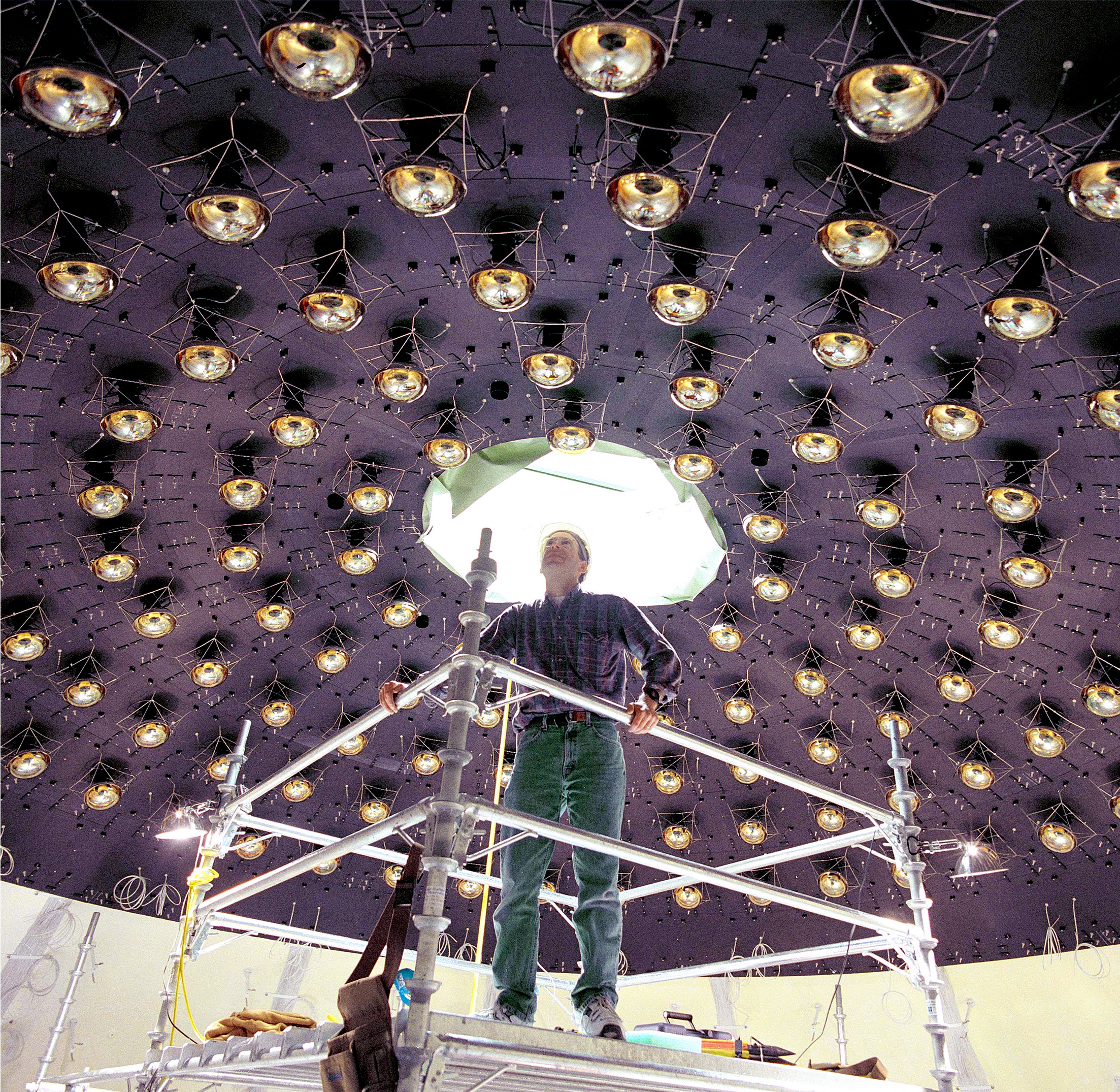 A man stands on a scaffold and inspects the ceiling of a large room, which is studded with reflective, semispherical detectors.