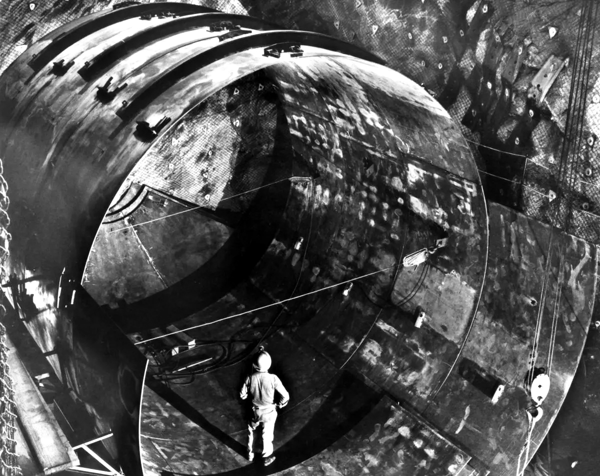 A man stands inside a metallic cylinder at least three times his height.