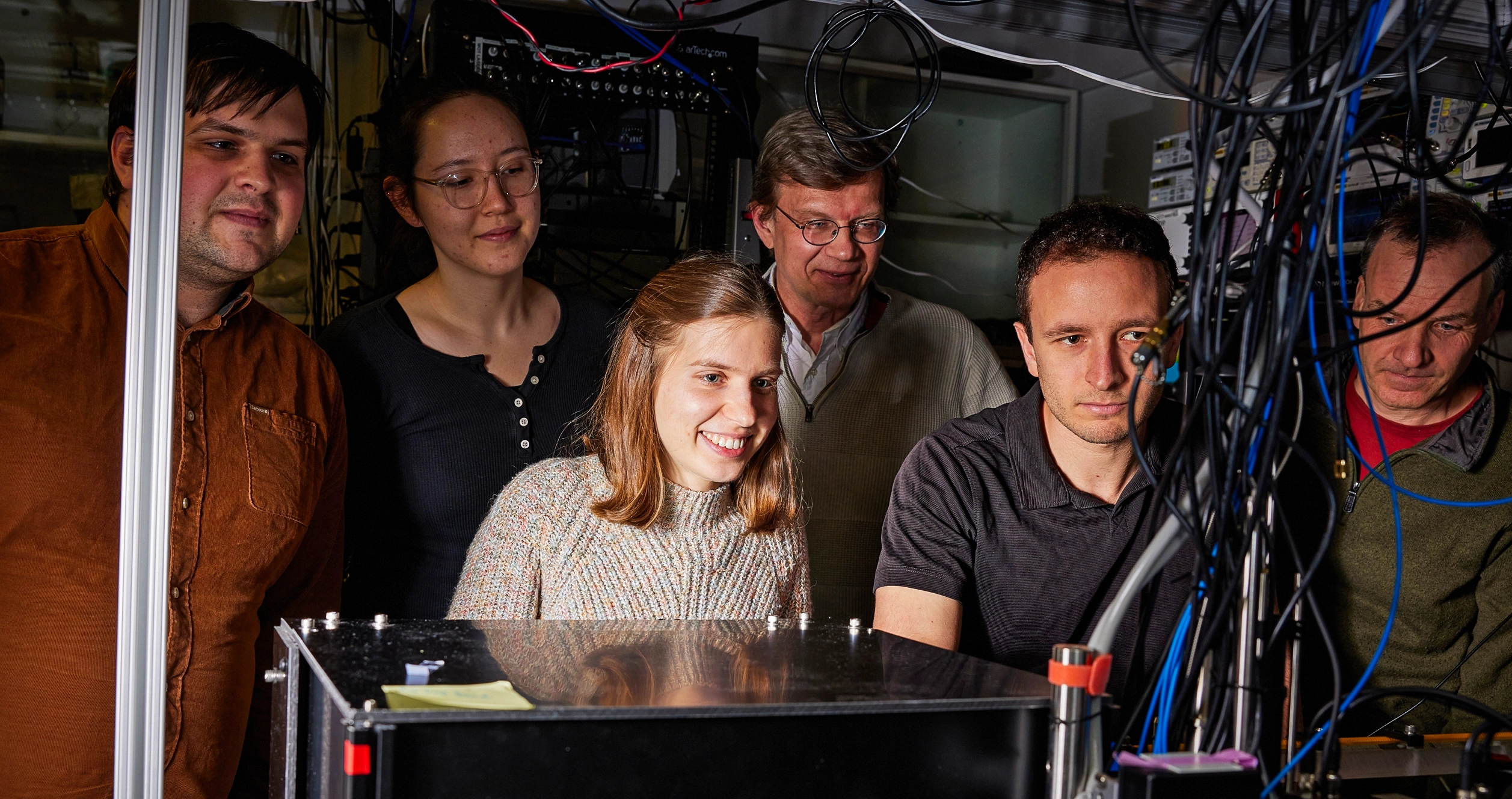 A group of six researchers gathered around laboratory equipment in a dimly lit lab filled with cables and electronic instruments.