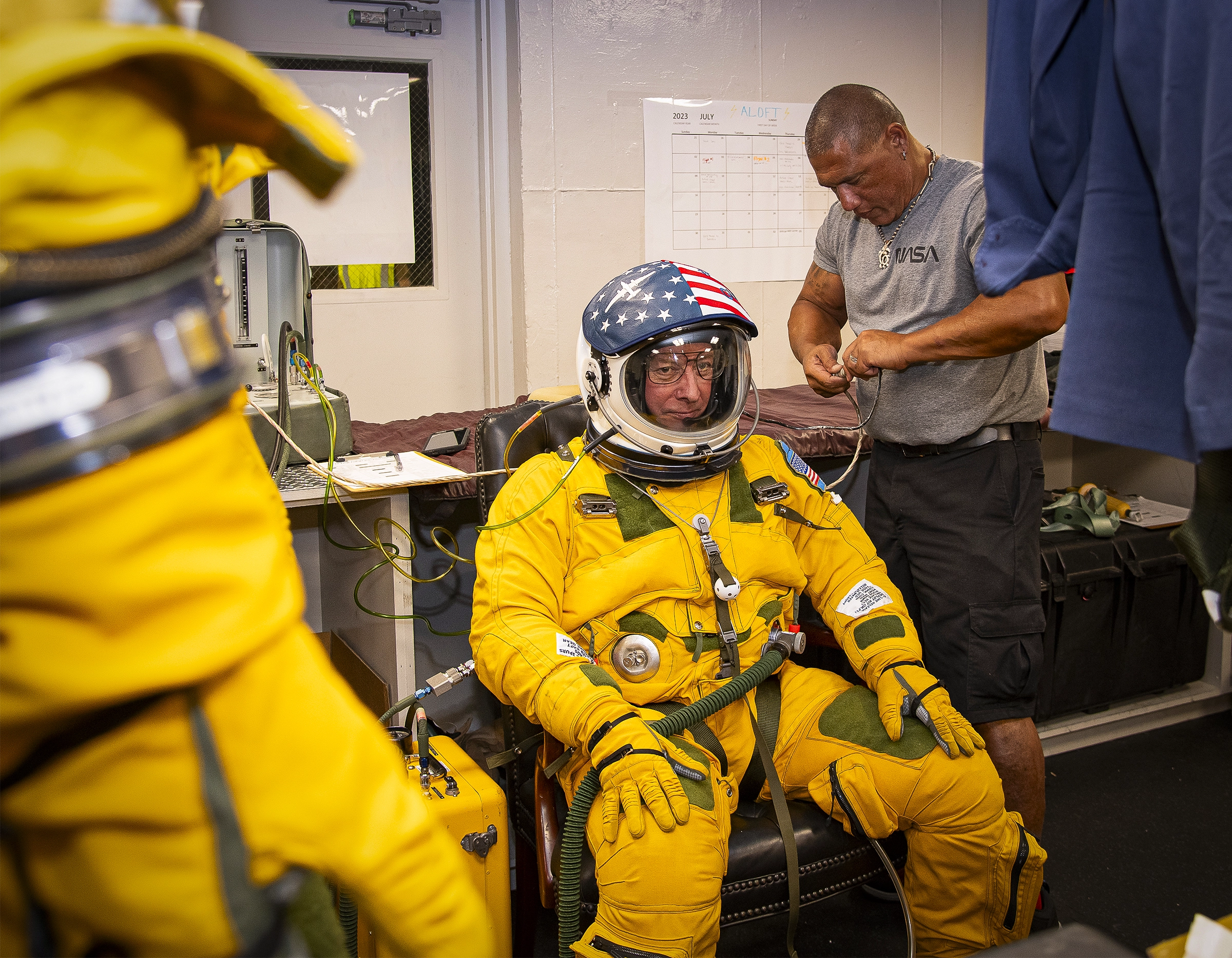 A pilot seated in a yellow high-altitude pressure suit with a stars-and-stripes helmet being assisted by a NASA technician in an equipment room before a flight.
