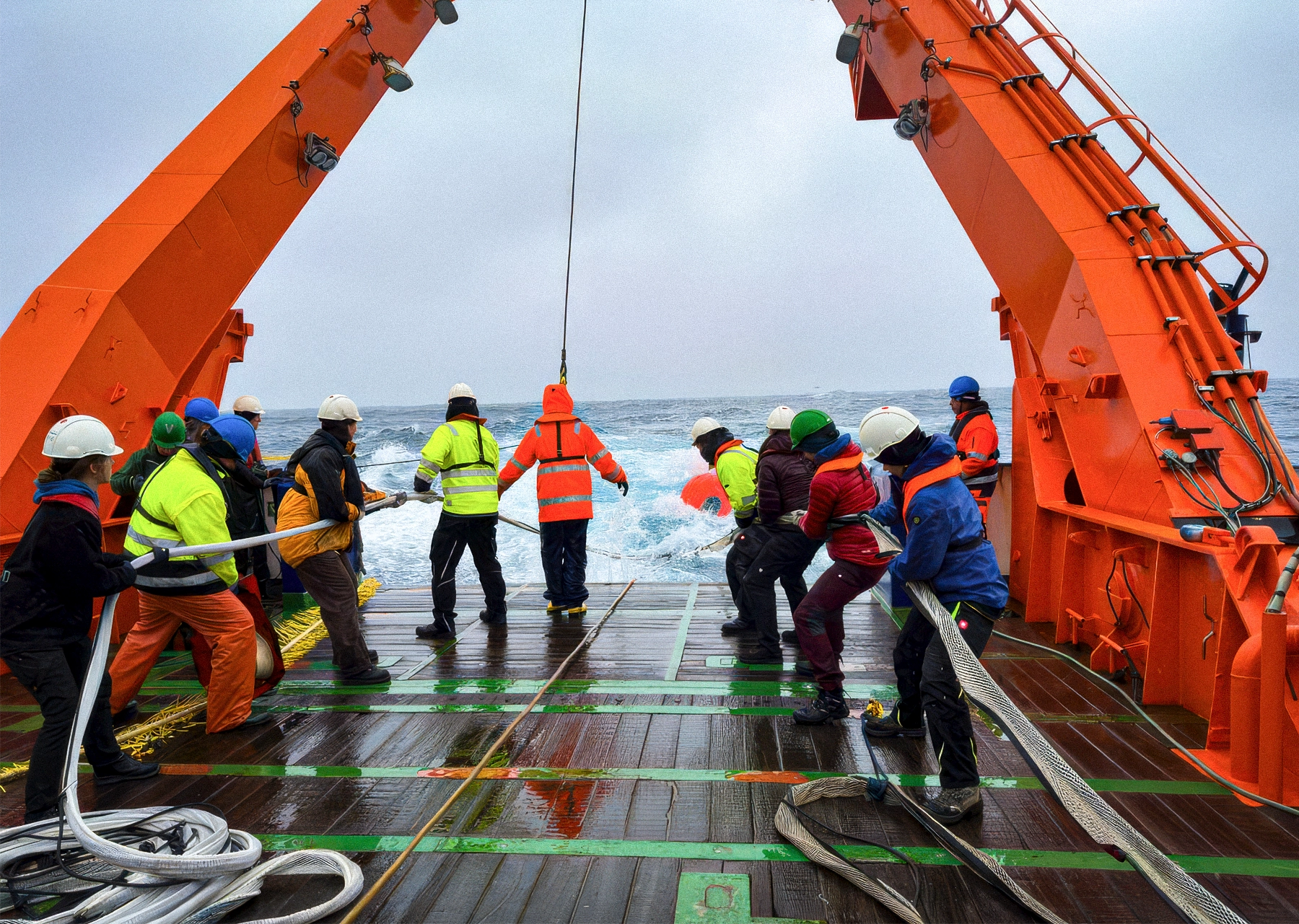 About a dozen people in boots and hard hats lower ropes into the ocean off the deck of a ship.