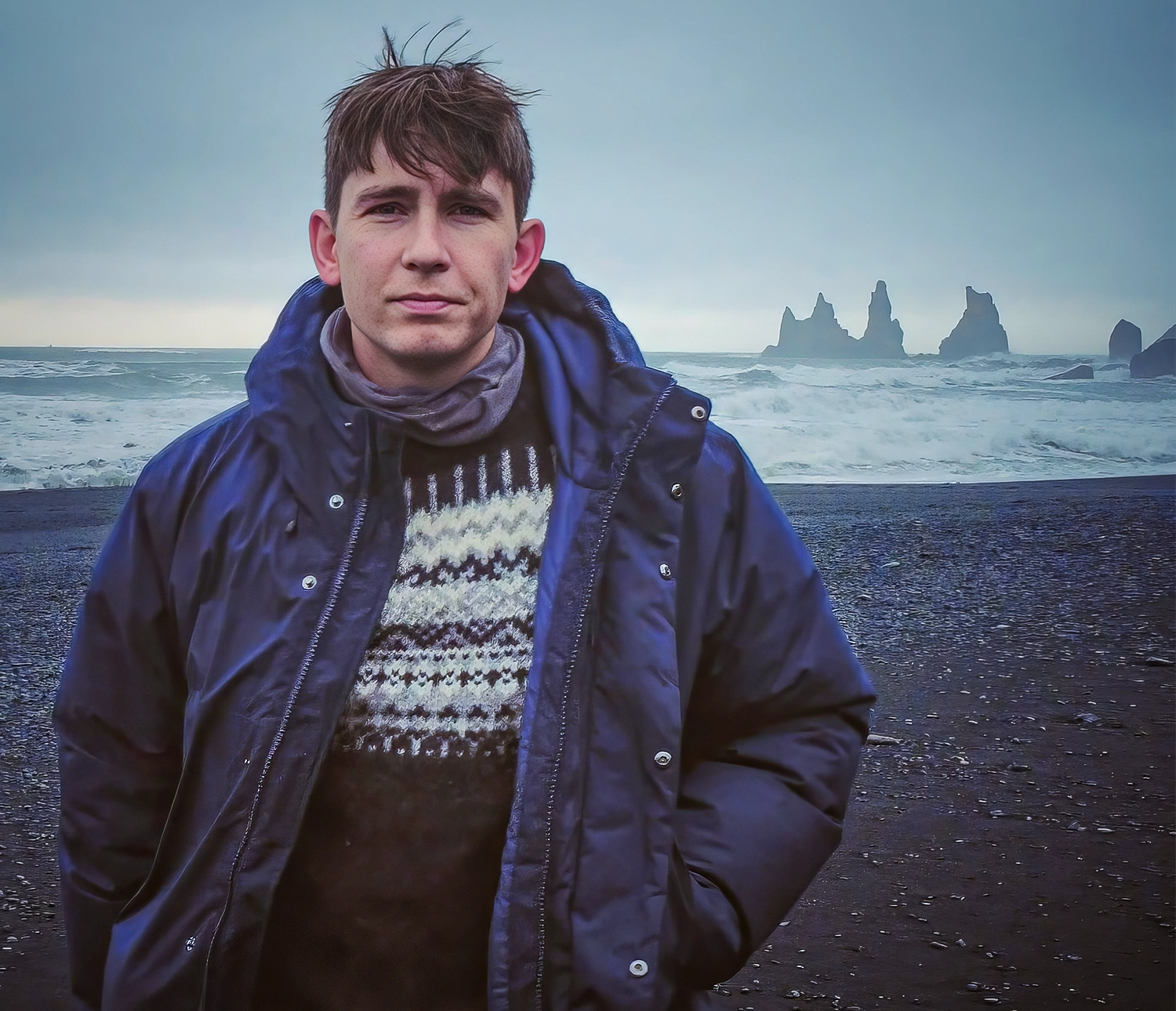 A man in a sweater and jacket stands on a beach in Iceland.