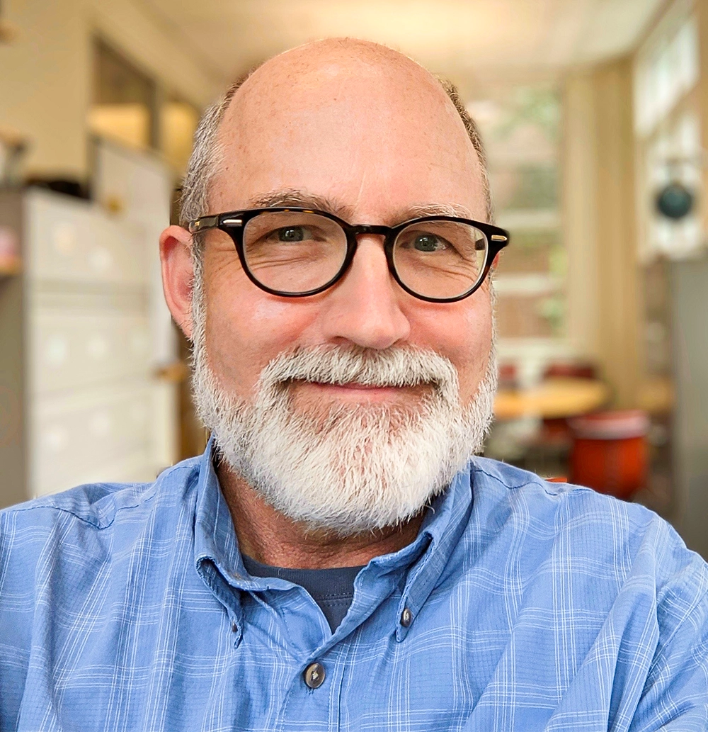 Headshot of a smiling older man with a gray beard and black-rimmed glasses, wearing a blue plaid shirt, photographed indoors against a softly blurred background.