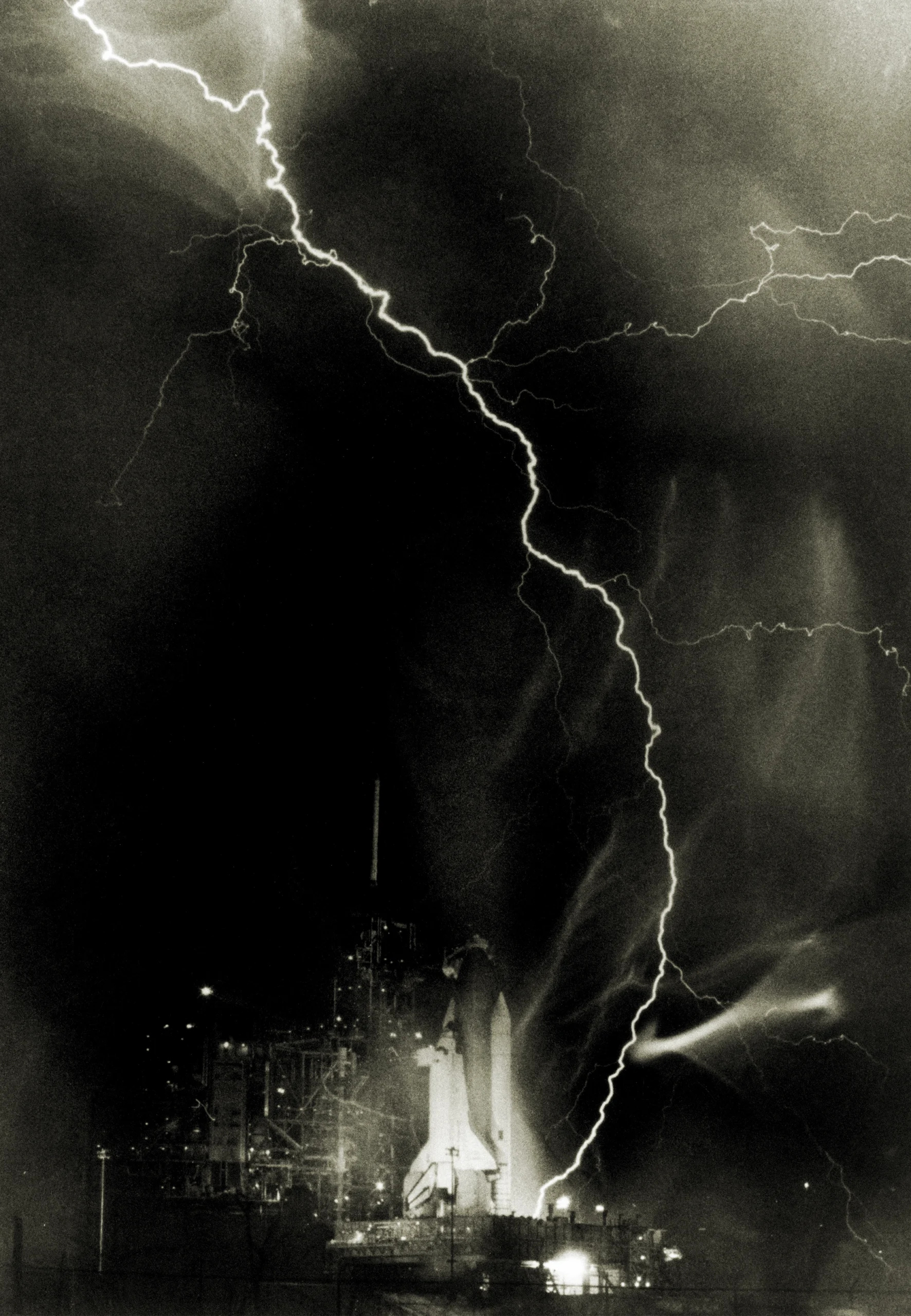 Black and white nighttime photograph of a space shuttle on its launchpad with a dramatic lightning bolt striking directly overhead against a dark stormy sky.