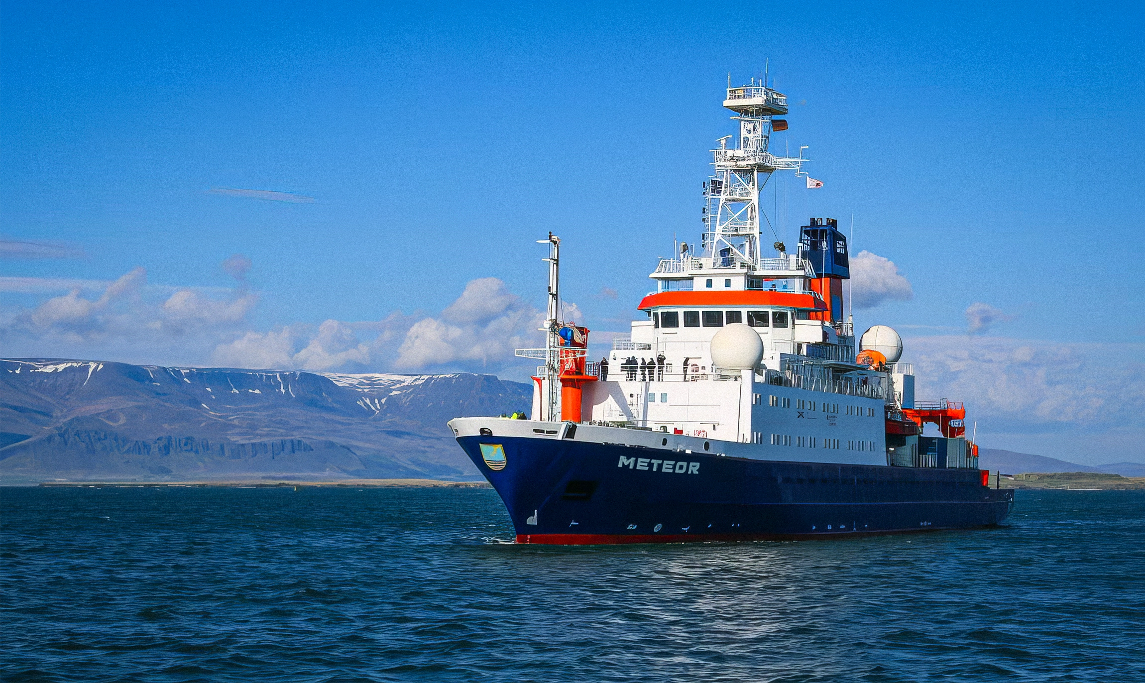 A boat about 100 meters in length sails on the ocean past an expanse of land.
