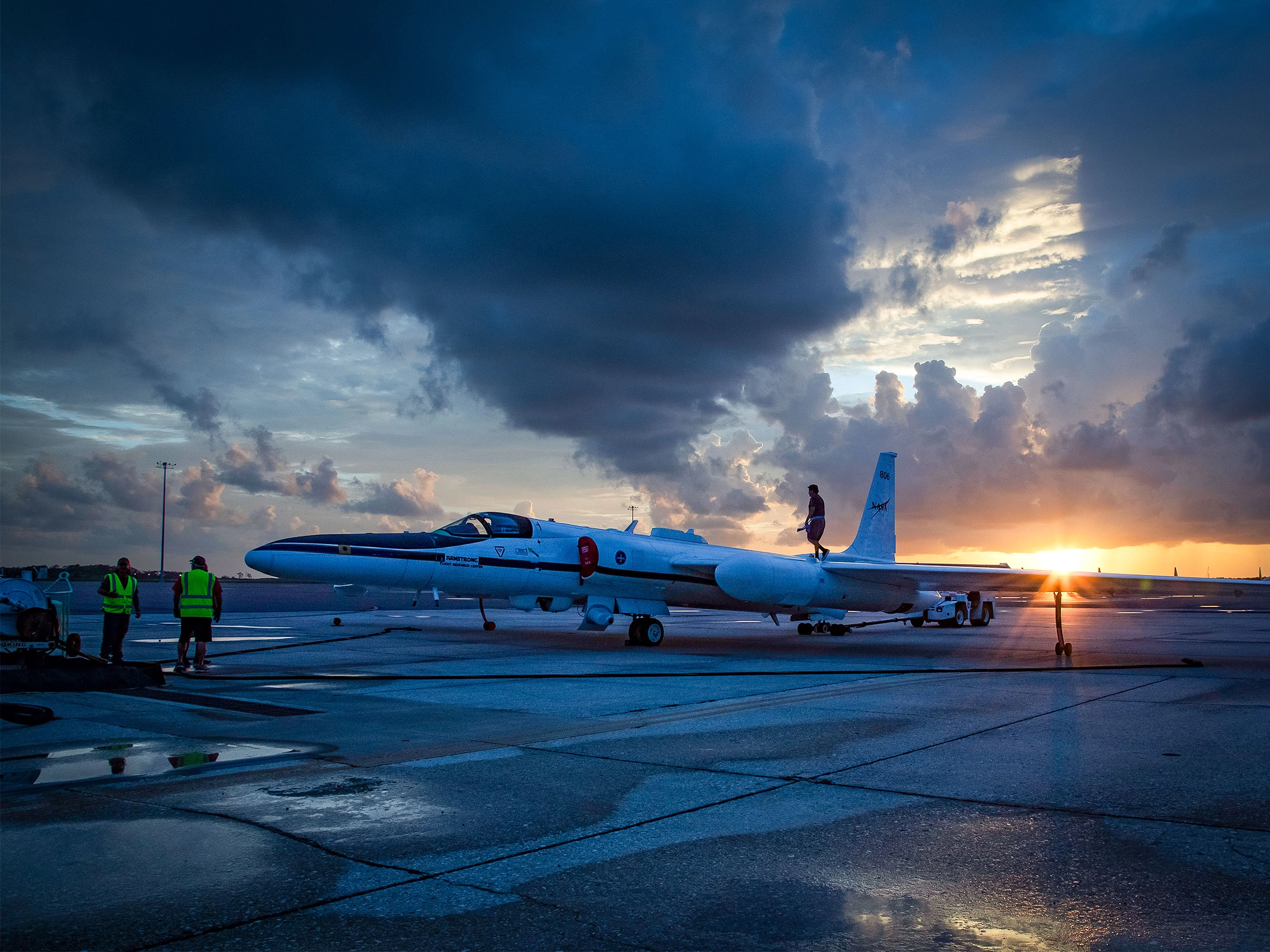 A NASA ER-2 high-altitude research aircraft on a wet runway at sunset, with ground crew in high-visibility vests visible nearby beneath a dramatic stormy sky.