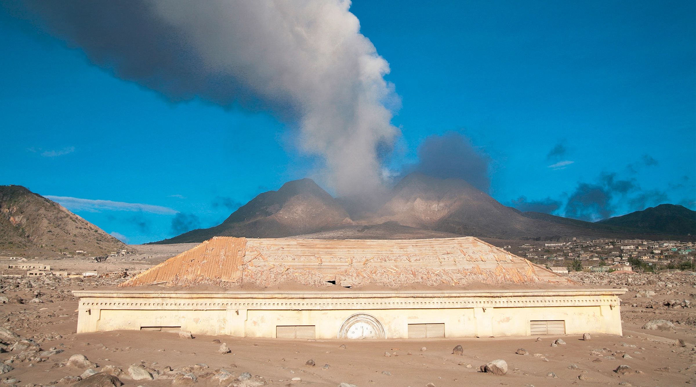 A smokey volcano behind a house submerged within sand and ash