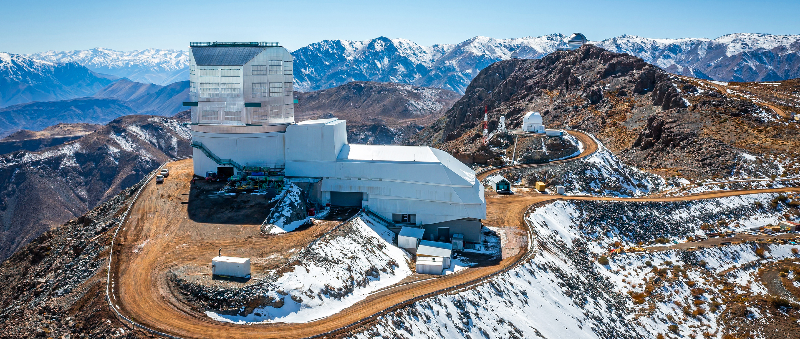 An approximately L-shaped white building sits on a peak surrounded by snowy mountains. A dirt road leads away from the building down the mountain.