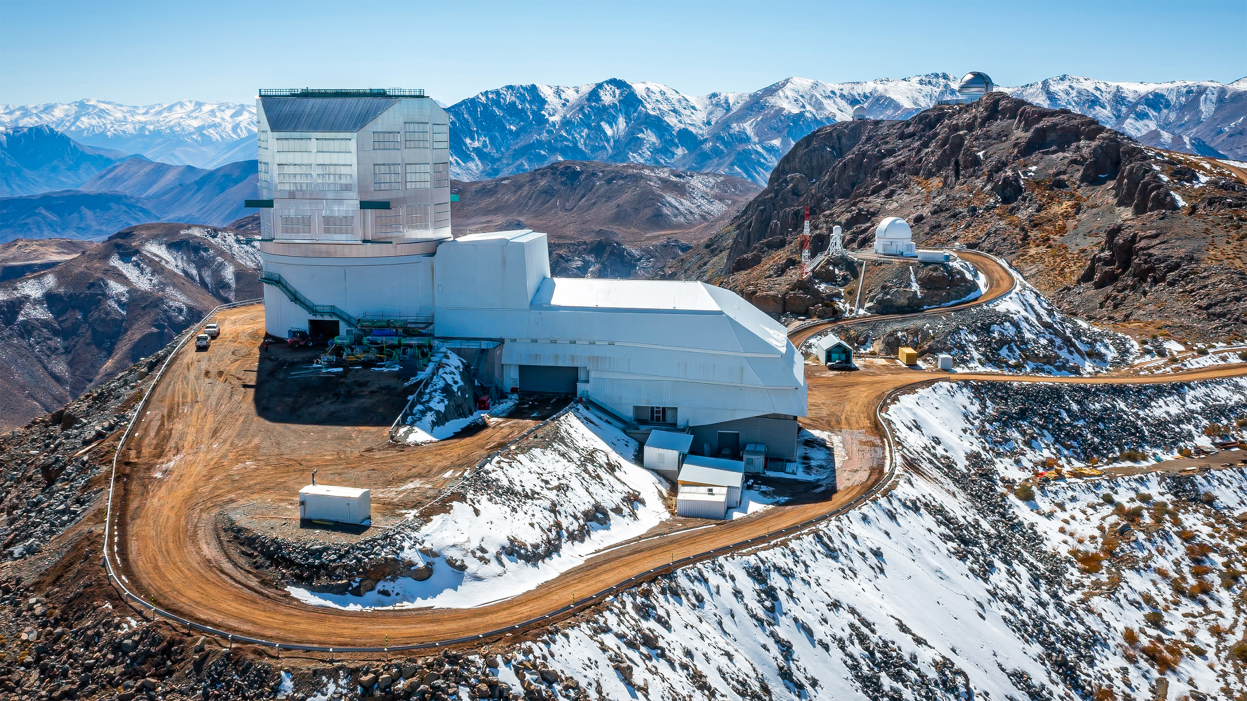 An approximately L-shaped white building sits on a peak surrounded by snowy mountains. A dirt road leads away from the building down the mountain.