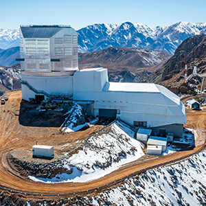 An approximately L-shaped white building sits on a peak surrounded by snowy mountains. A dirt road leads away from the building down the mountain.