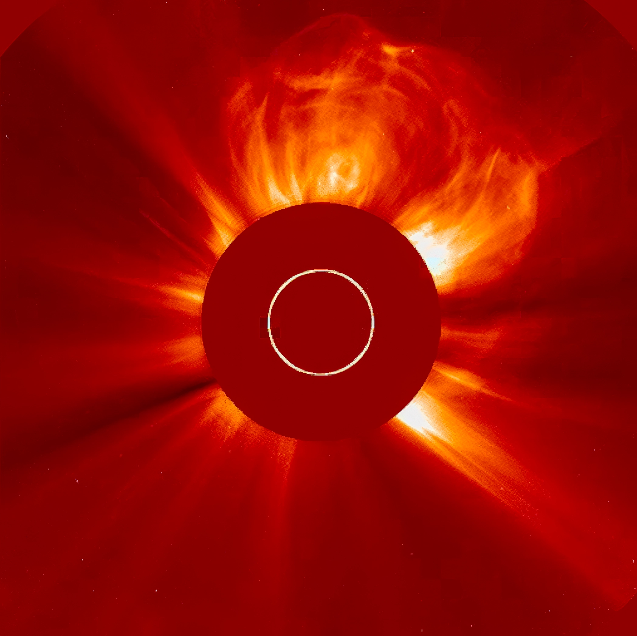 A coronagraph image of the sun in false-color red and orange. The sun’s bright disk is blocked by a circular mark at the center.