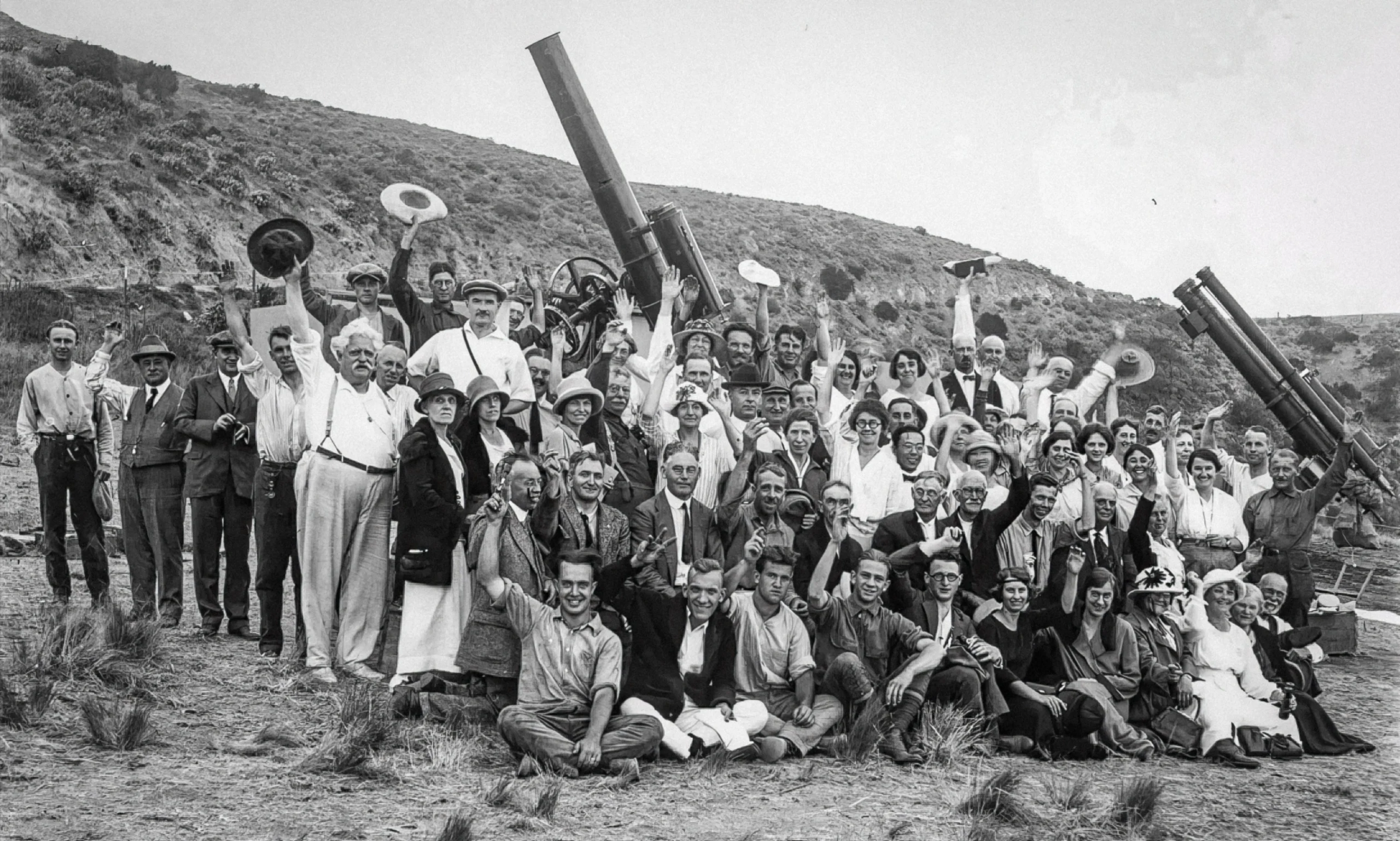 A black-and-white photograph from the early 20th century showing a large group of approximately 50 people posing outdoors on a hillside around two telescopes. The group includes men and women arranged in several rows, with some seated on the ground in front and others standing behind. Some people are waving hats and raising their arms in celebration.
