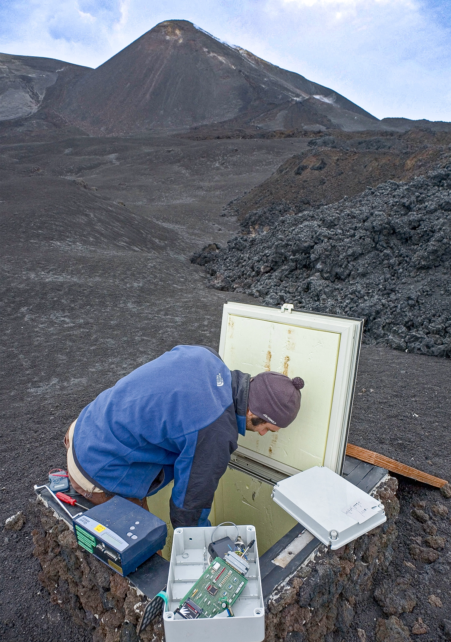 A volcanologist reaching into a compartment embedded in the ground near a volcano