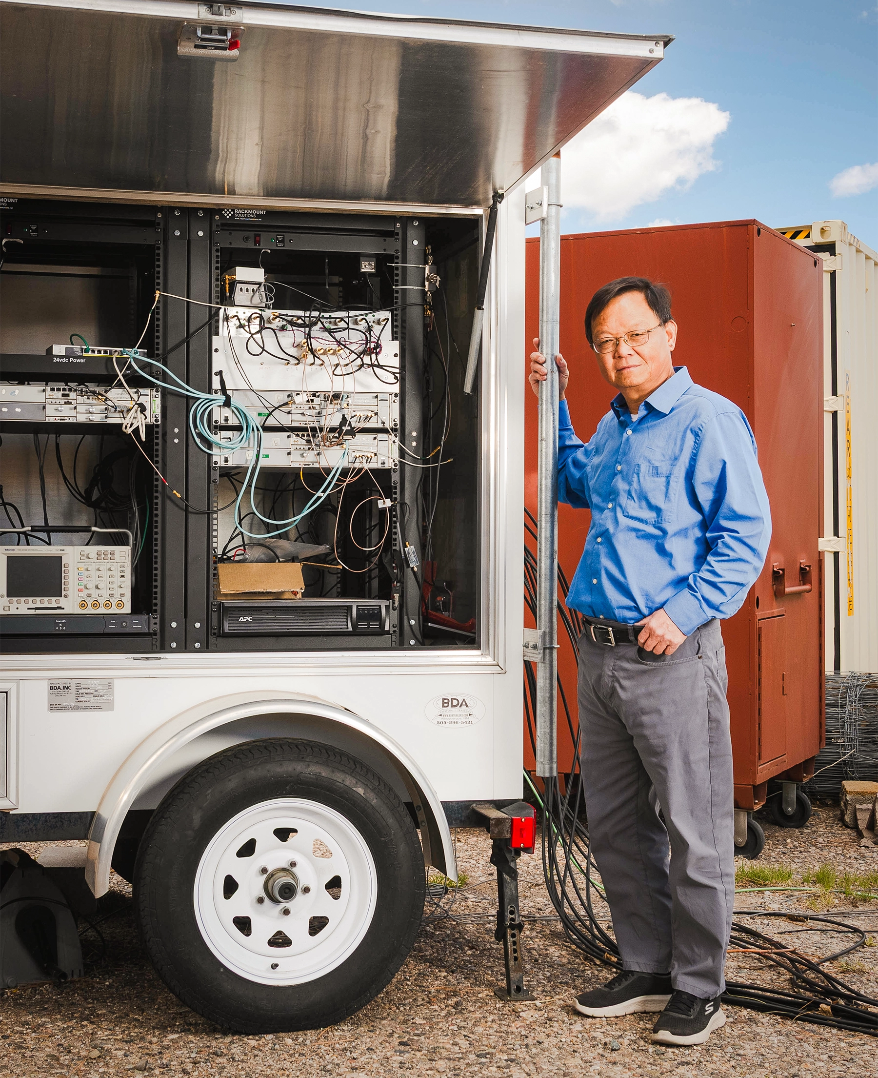 Photograph of an older male researcher standing beside a mobile scientific instrument trailer packed with electronic equipment, cables, and hardware. They are outdoors on a gravel surface under a blue and cloudy sky.