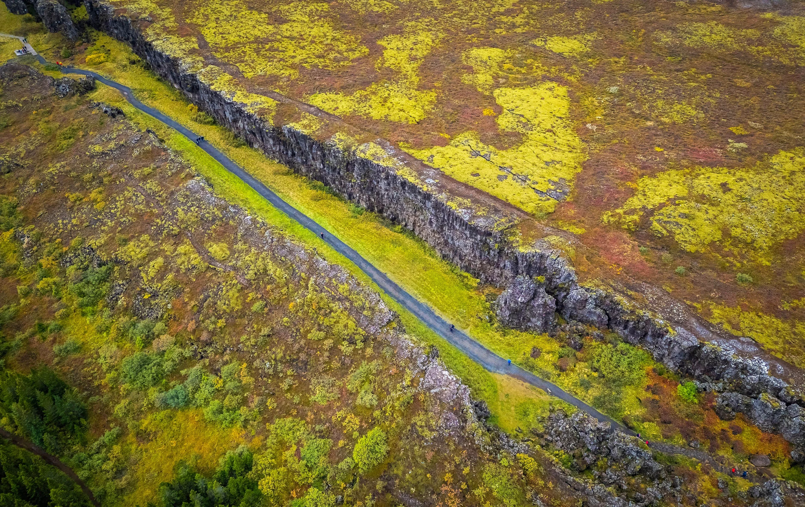An aerial view shows a few hikers walking down a paved path bracketed by two walls of rock.