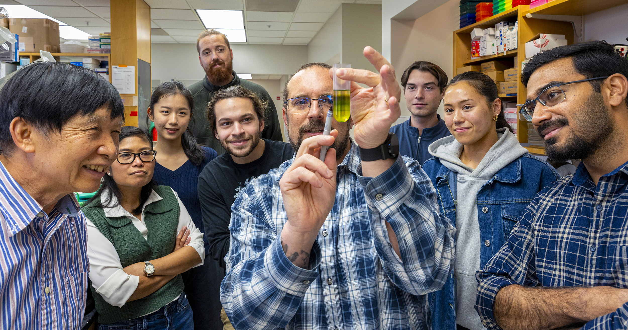 Chris Gisriel, surrounded by eight colleagues, holds a flask of cyanobacteria.