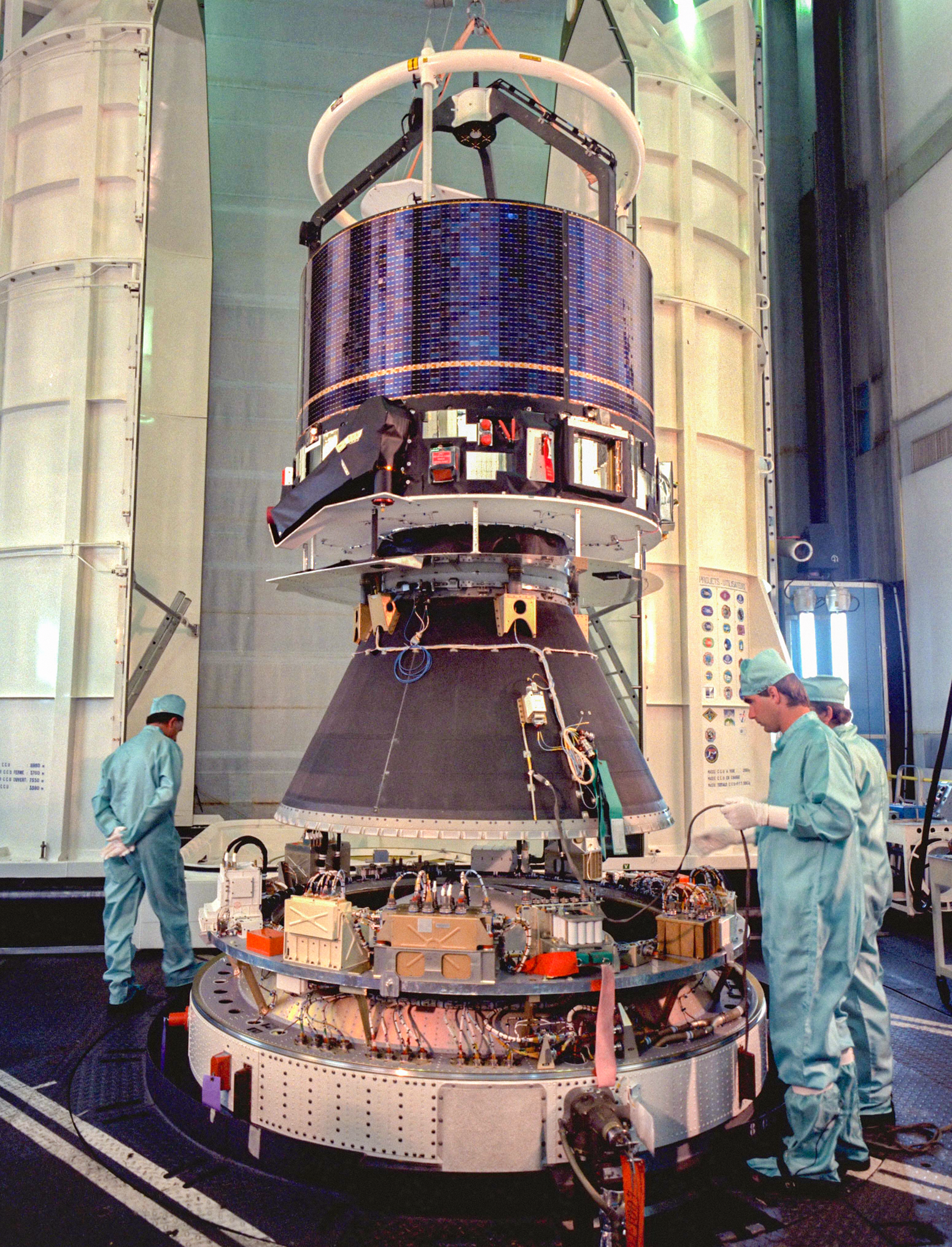Photograph of the Giotto spacecraft under construction in a tall indoor space with three people in clean room suits.