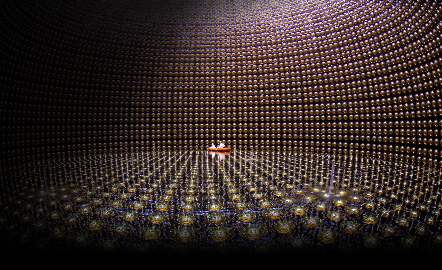 A large cavern lined with reflective spheres and partially filled with water. Two people in white protective gear float in an orange raft in the distance.