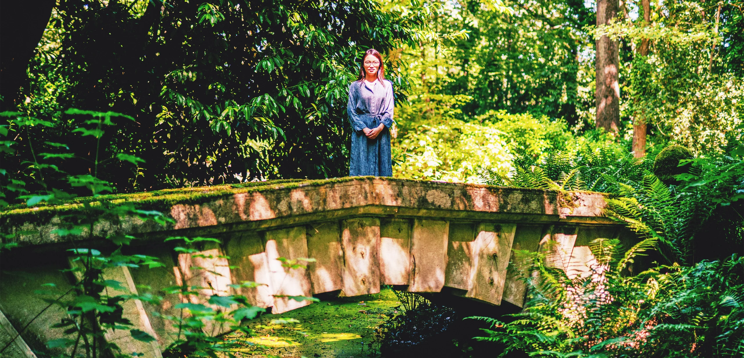A woman in a blue dress stands on a low, stone bridge, surrounded by greenery.