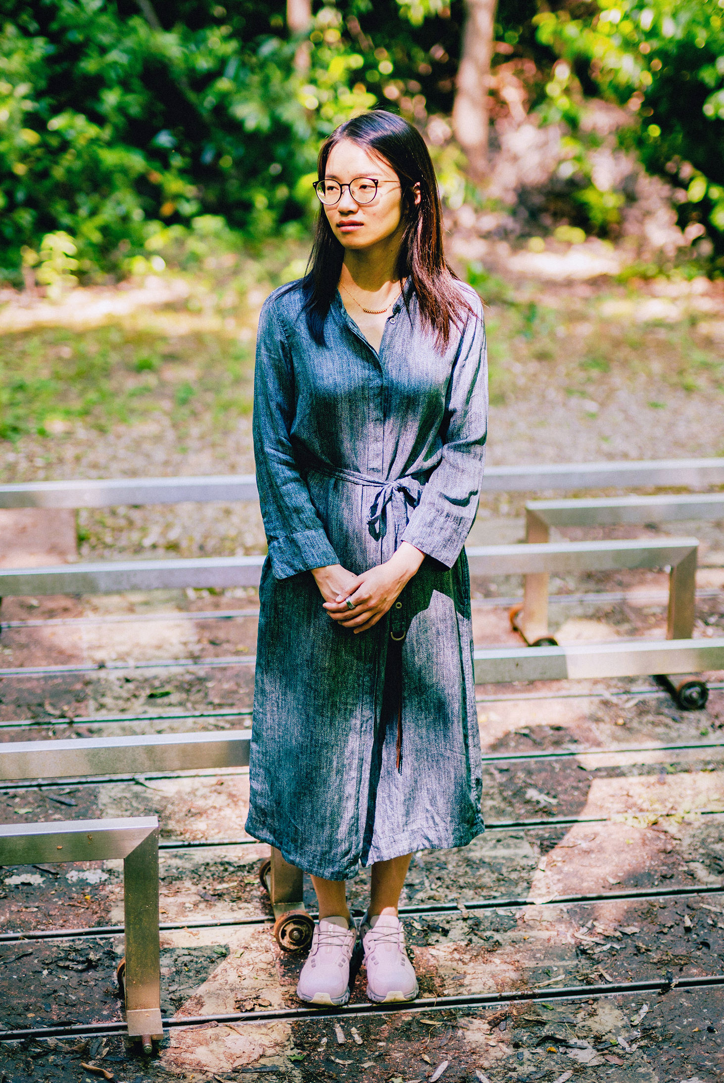 A woman in a blue dress stands in front of a low, angular sculpture near some trees.