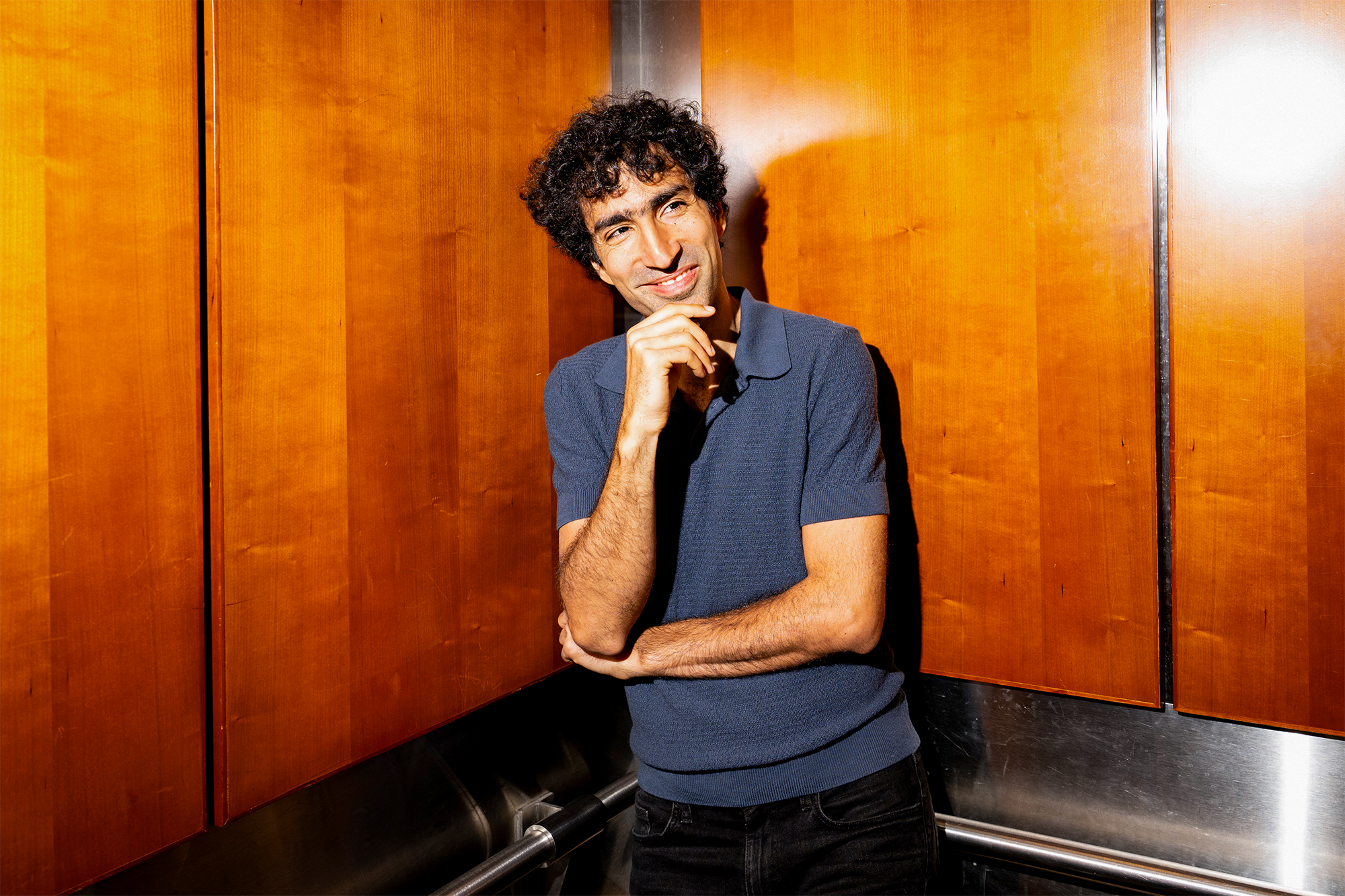 Shayan Oveis Gharan, wearing a navy polo shirt, stands smiling in a wood-paneled elevator with his arms crossed and one hand resting thoughtfully on his chin.