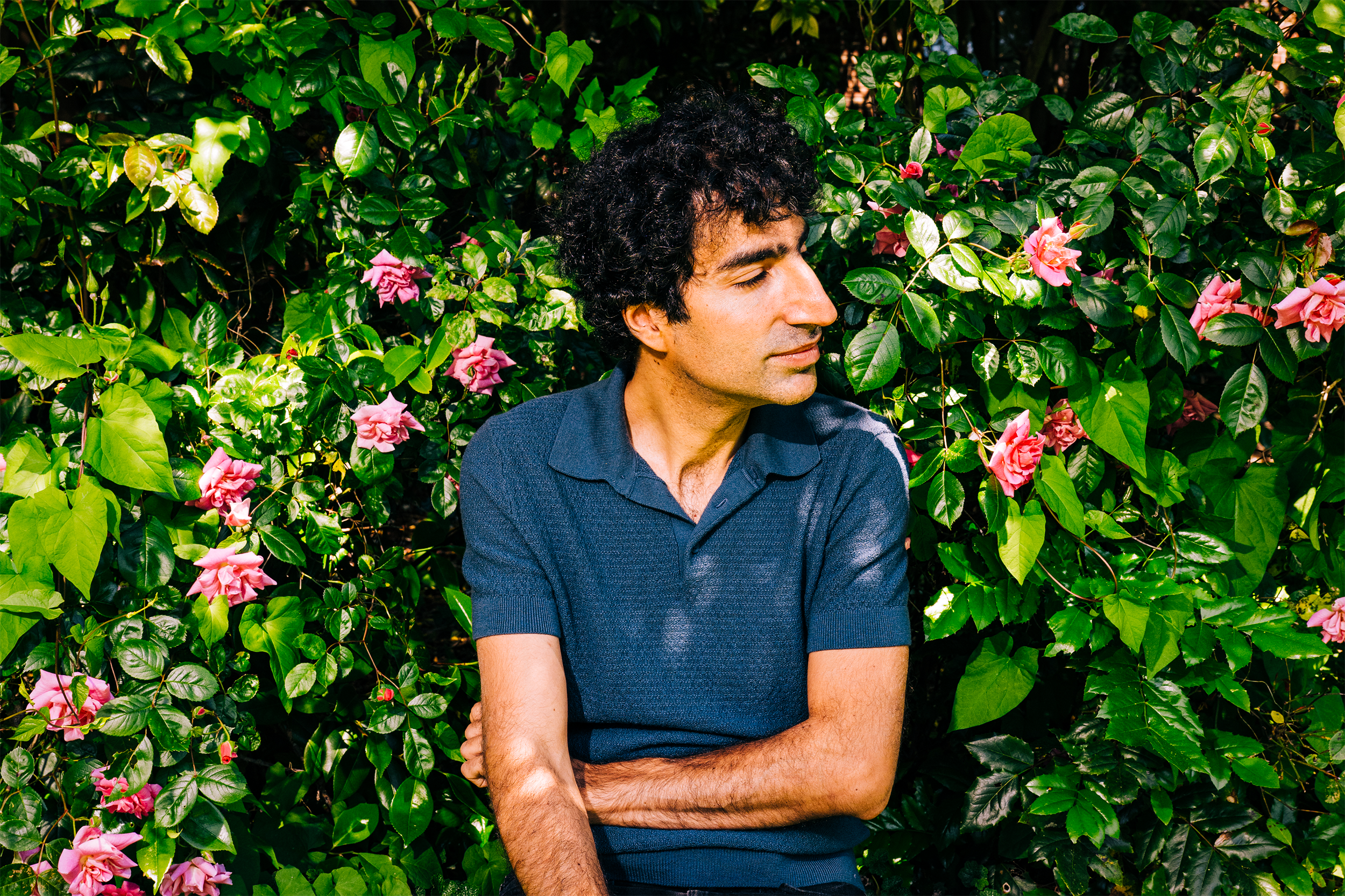 Shayan Oveis Gharan with his arms partially crossed and eyes closed in front of a rose bush, his face turned to one side.