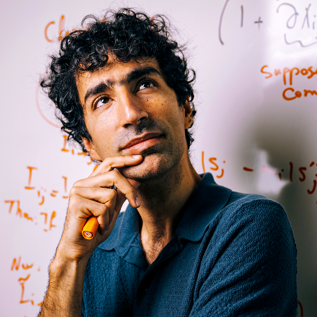 Shayan Oveis Gharan holds a marker in his hand up to his chin, and looks upward in thought in front of a whiteboard covered in mathematical notation.