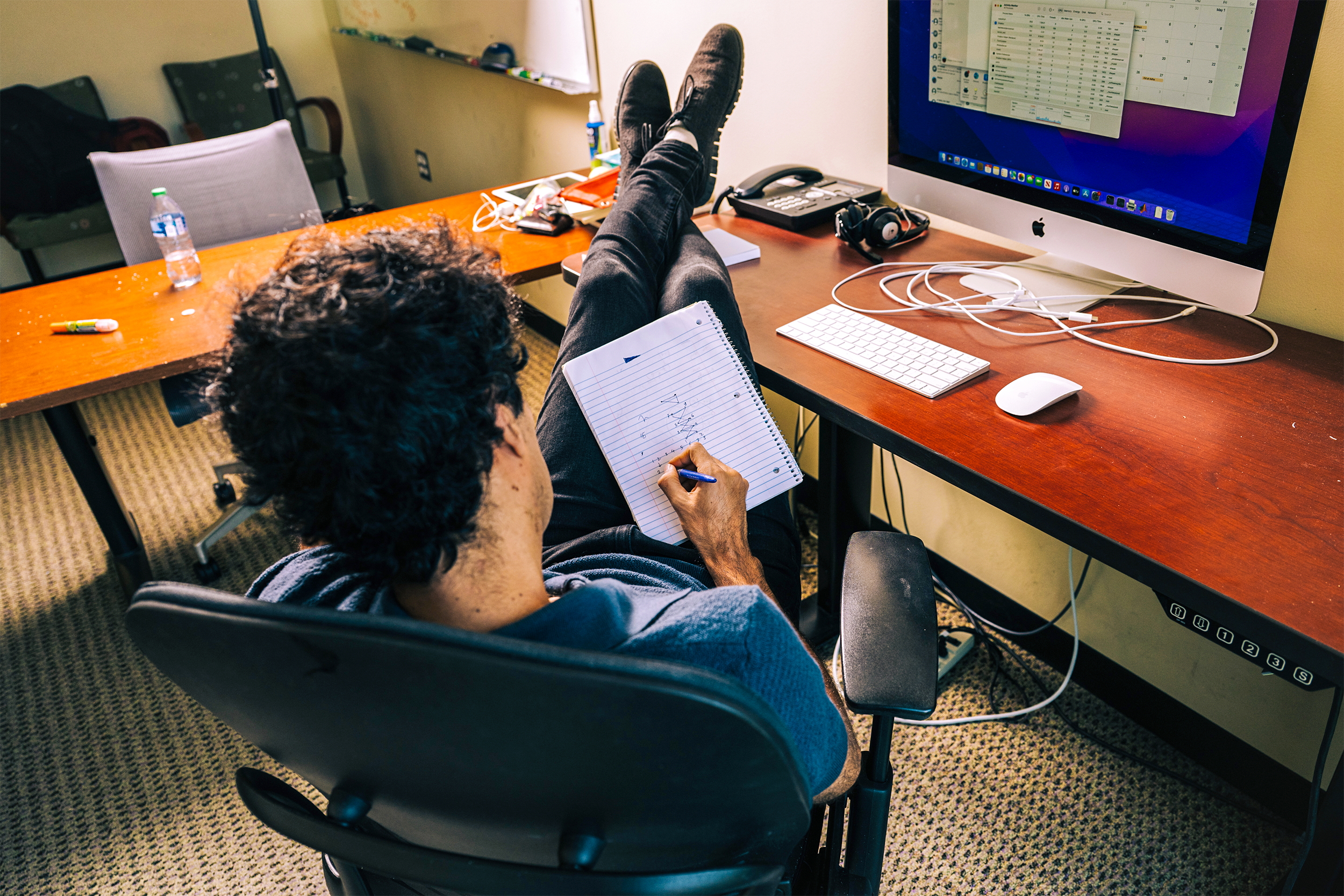 Shayan Oveis Gharan reclines in an office chair with his feet up on the desk in front of his computer, sketching a graph diagram on a notepad.