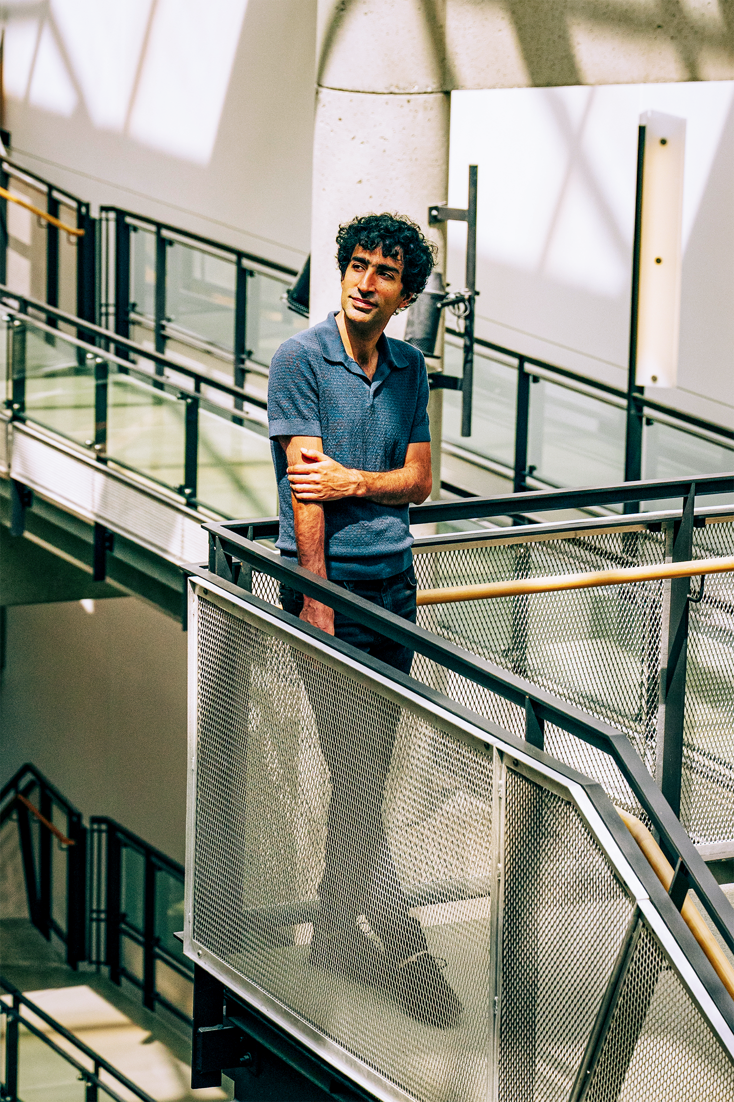 Shayan Oveis Gharan stands on a metal staircase landing in a modern building, arms crossed, gazing thoughtfully upward and to the side.