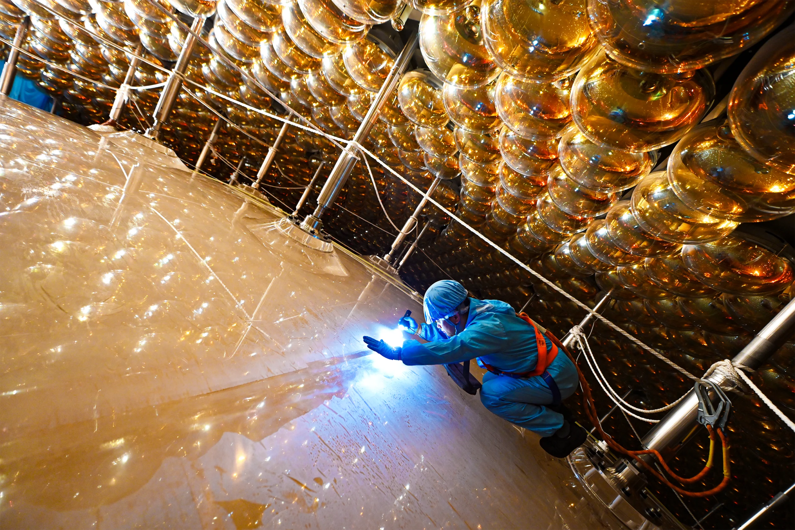 A man in both clean-room and climbing gear shines a flashlight while bracing himself against a surface inside the JUNO detector.