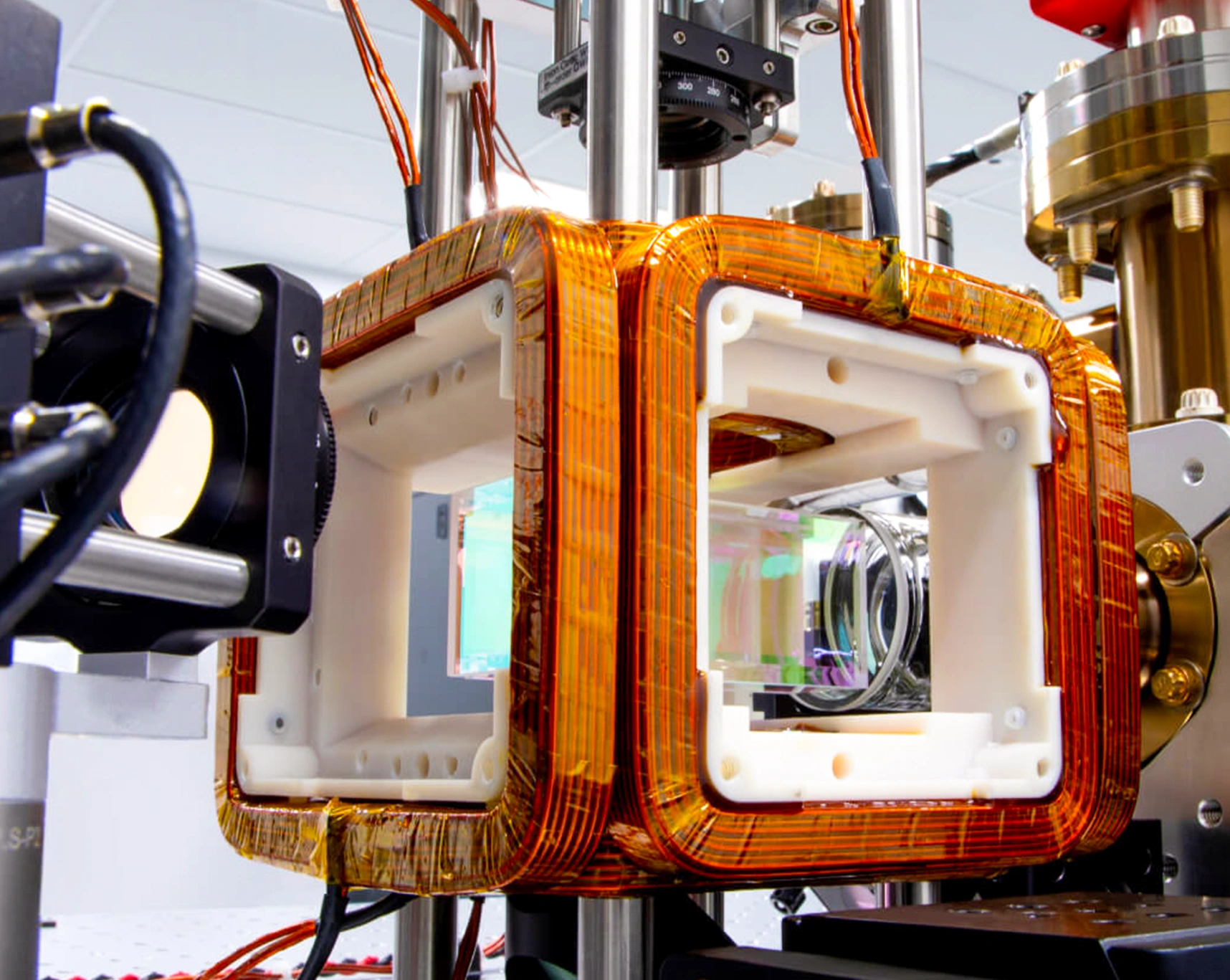 A glass vacuum cell surrounded by a white housing unit wound with copper wire coils, surrounded by components used to focus laser beams into the cell.
