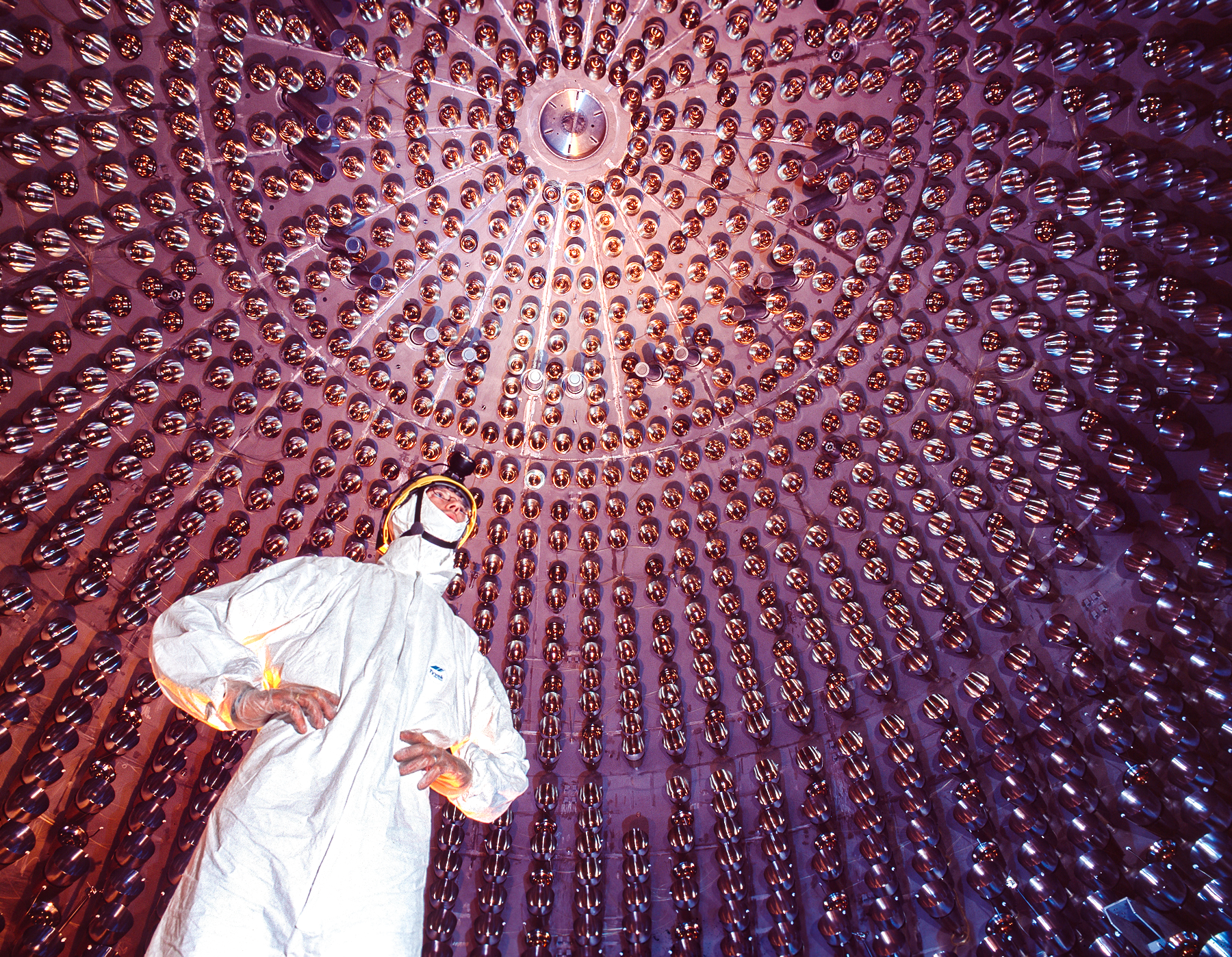 A man in a white bunny suit and hard hat stands inside a giant spherical detector lined with photomultiplers that resemble large, gold-tinted lightbulbs.