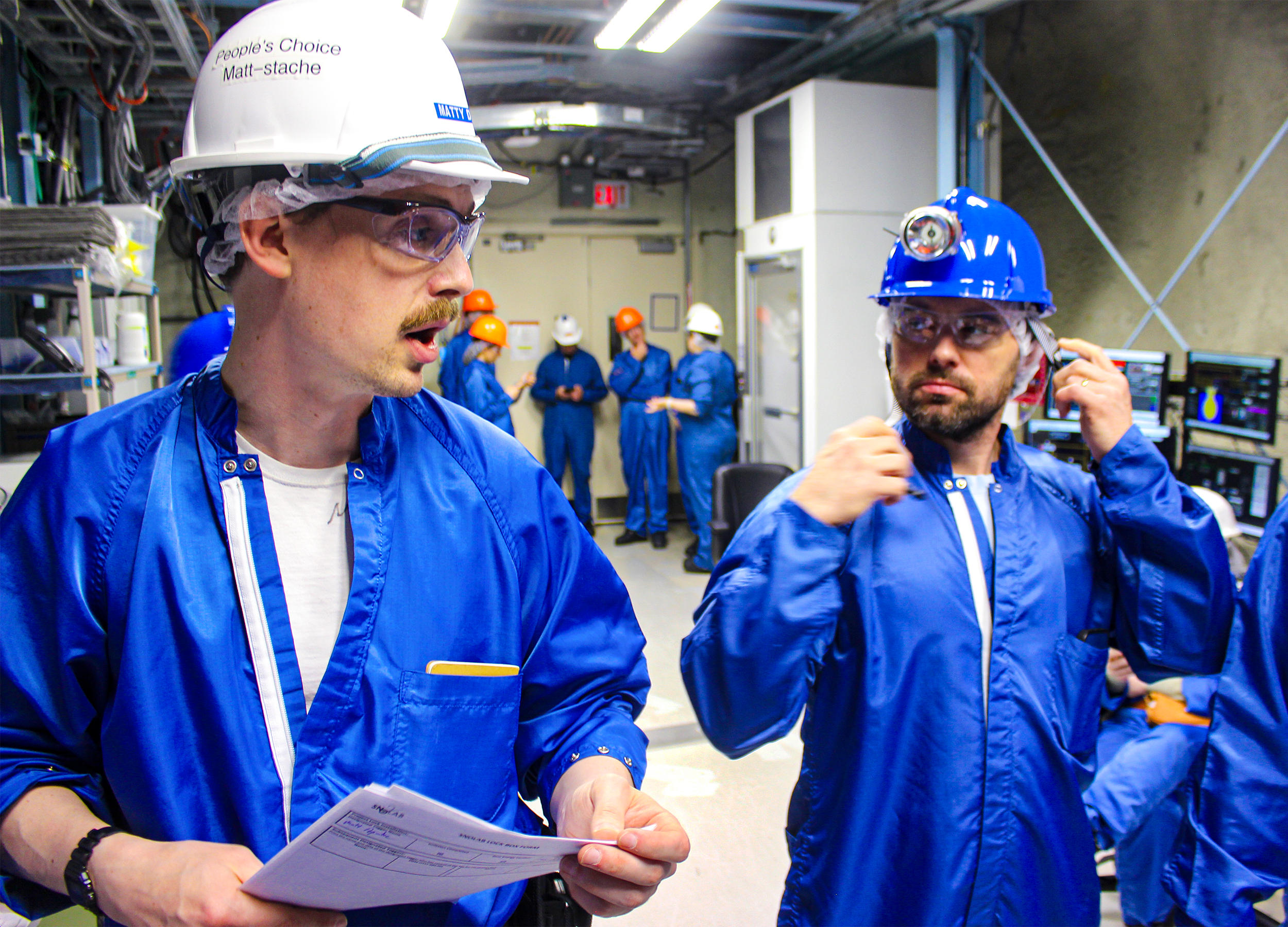 A mustachioed man in a blue jumpsuit and a white hardhat, which is labeled with both his name and the text “People’s Choice Matt-stache.” He is walking with others in a well-lit area of an underground laboratory.