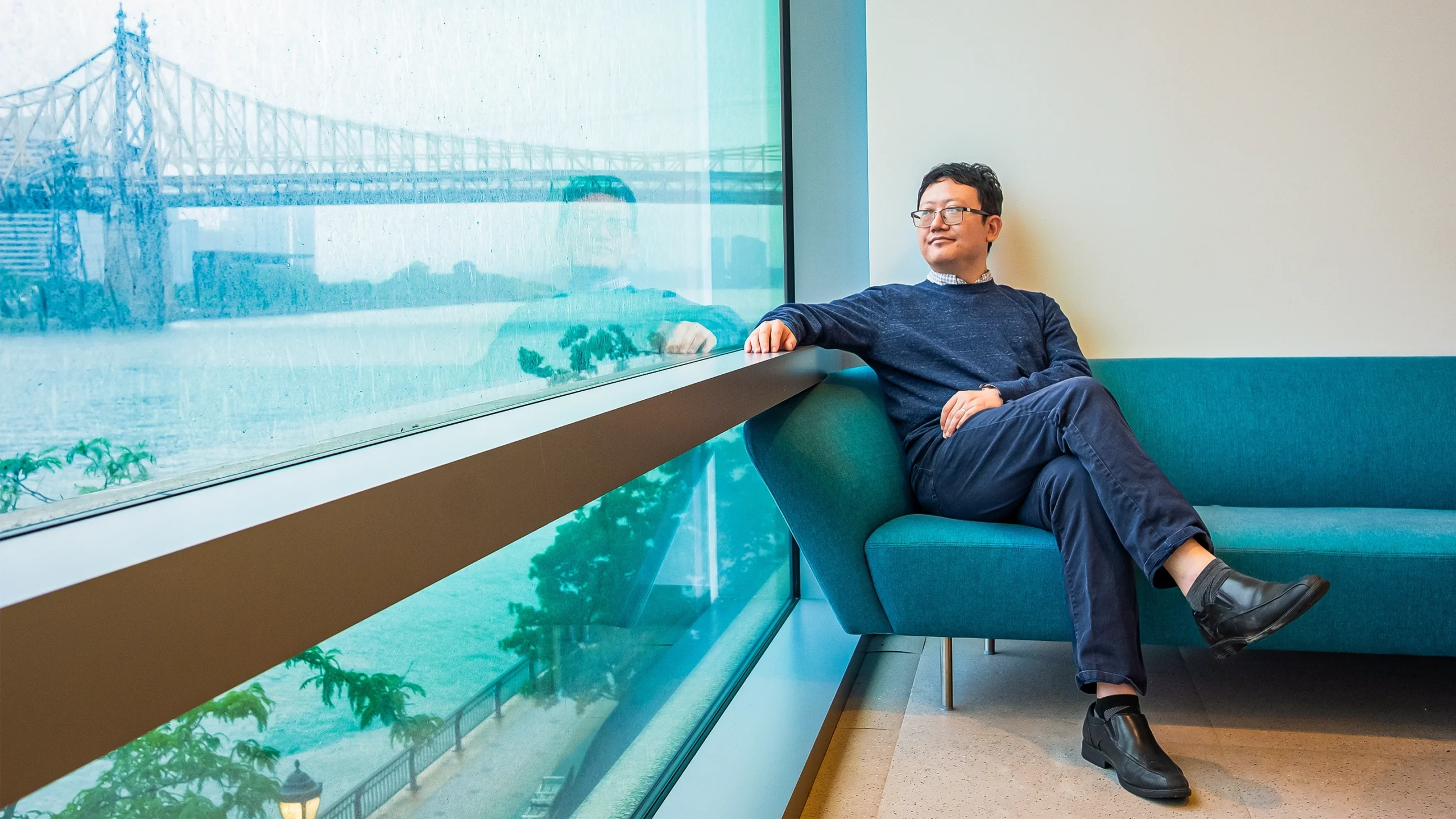 A man in a sweater sitting on a blue couch, looking out of a window that reflects his image