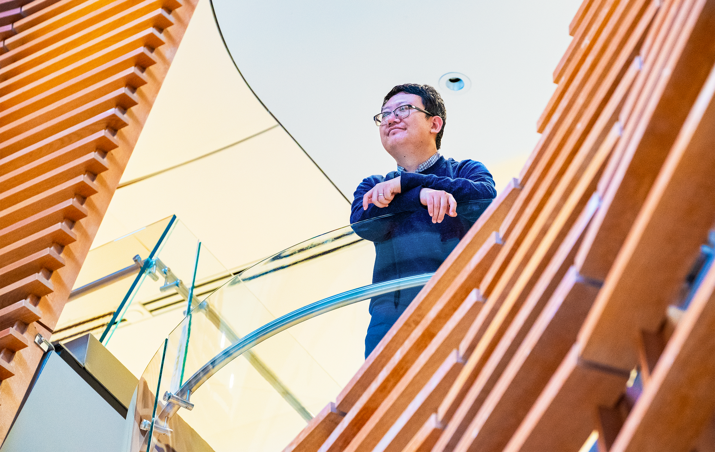 A man in a sweater standing on an indoor balcony looking out and smiling
