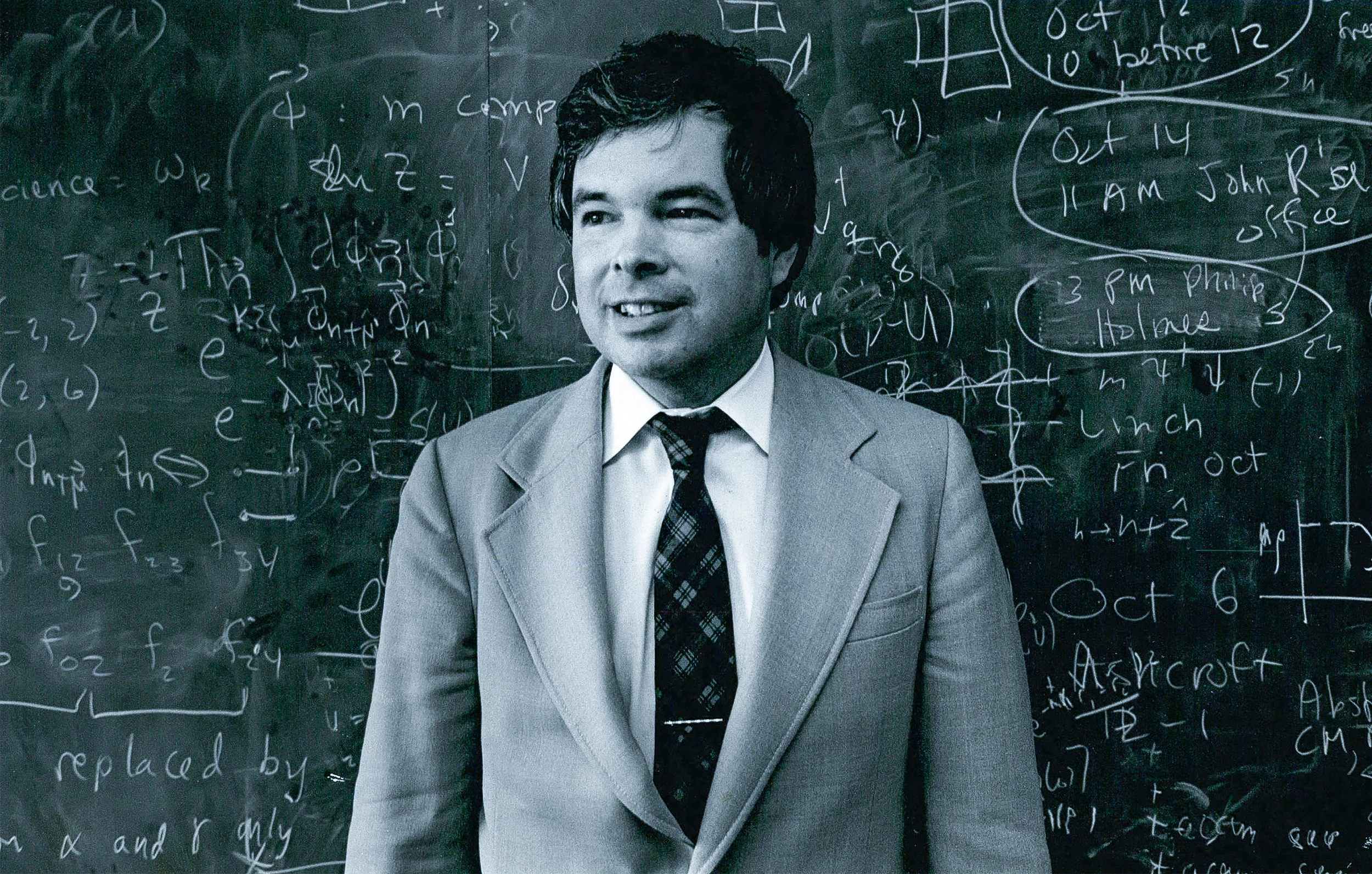 In a black-and-white photo, a man in a suit stands in front of a chalkboard.