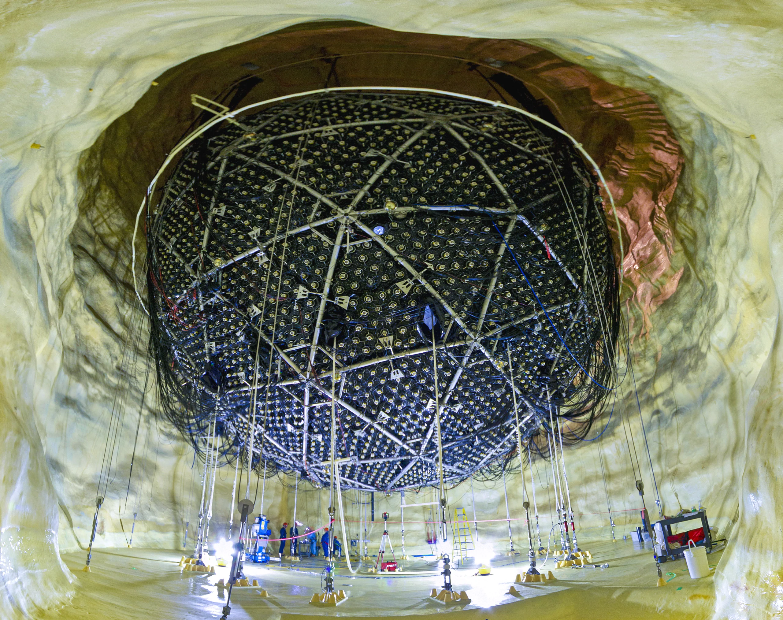 People in blue bunny suits and hard hats stand beneath a giant spherical particle detector in a cavern with smooth white walls.