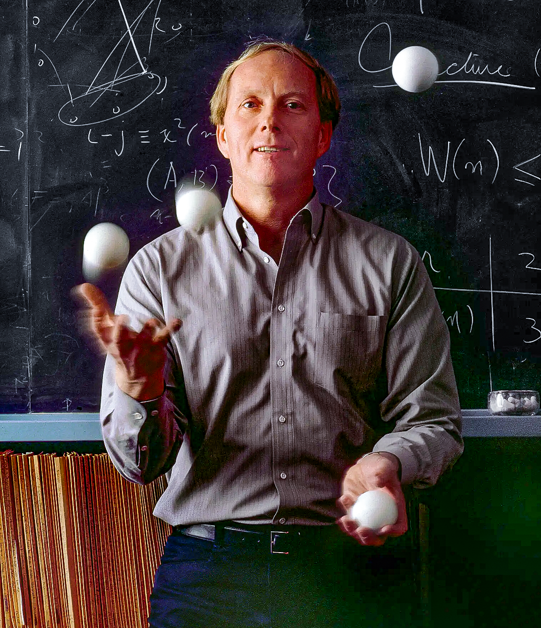 Man juggling four balls in front of a chalkboard.