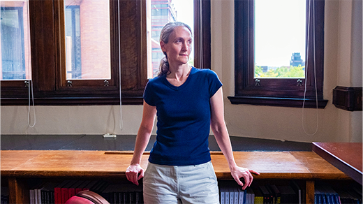 Demers, a woman with gray hair, stands in front of a window, leaning against a desk, looking out of frame.