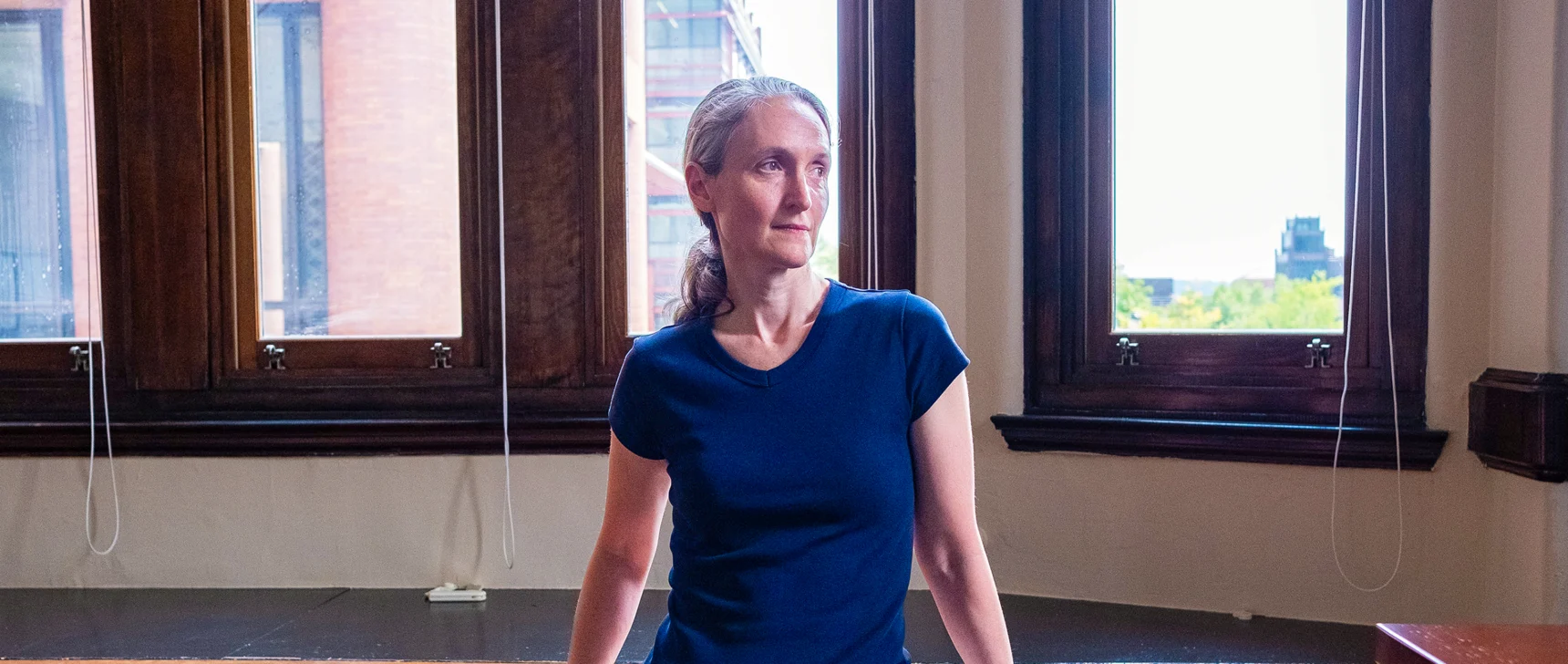 Demers, a woman with gray hair, stands in front of a window, leaning against a desk, looking out of frame.