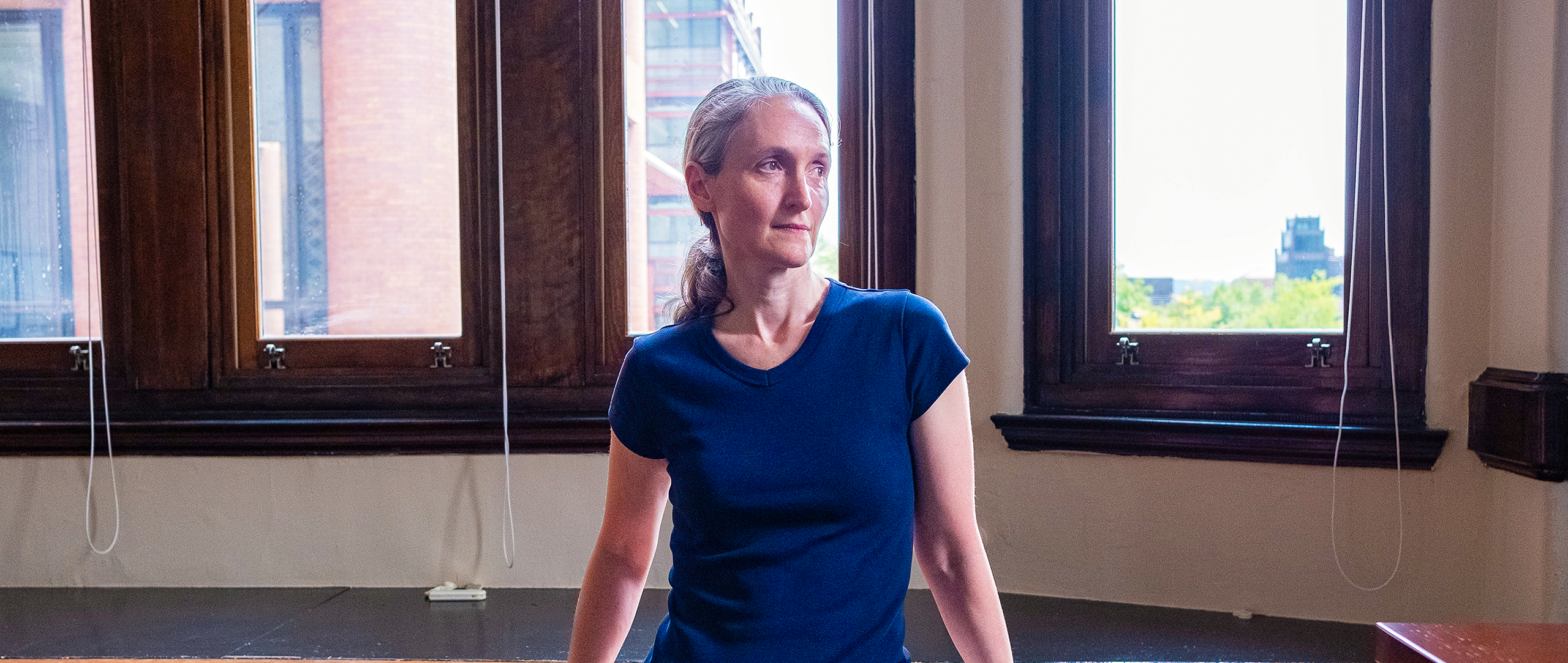 Demers, a woman with gray hair, stands in front of a window, leaning against a desk, looking out of frame.