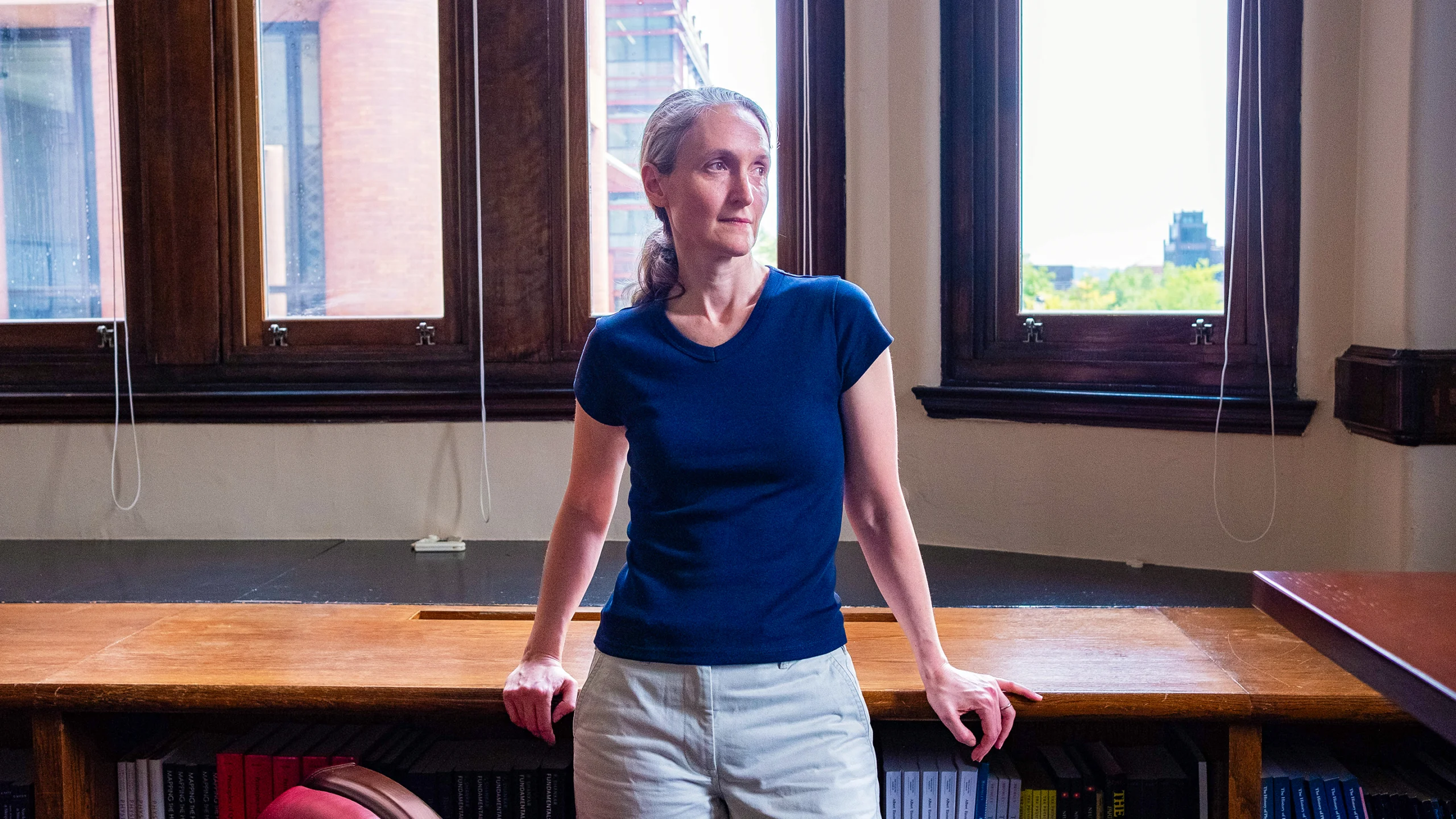 Demers, a woman with gray hair, stands in front of a window, leaning against a desk, looking out of frame.