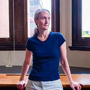 Demers, a woman with gray hair, stands in front of a window, leaning against a desk, looking out of frame.